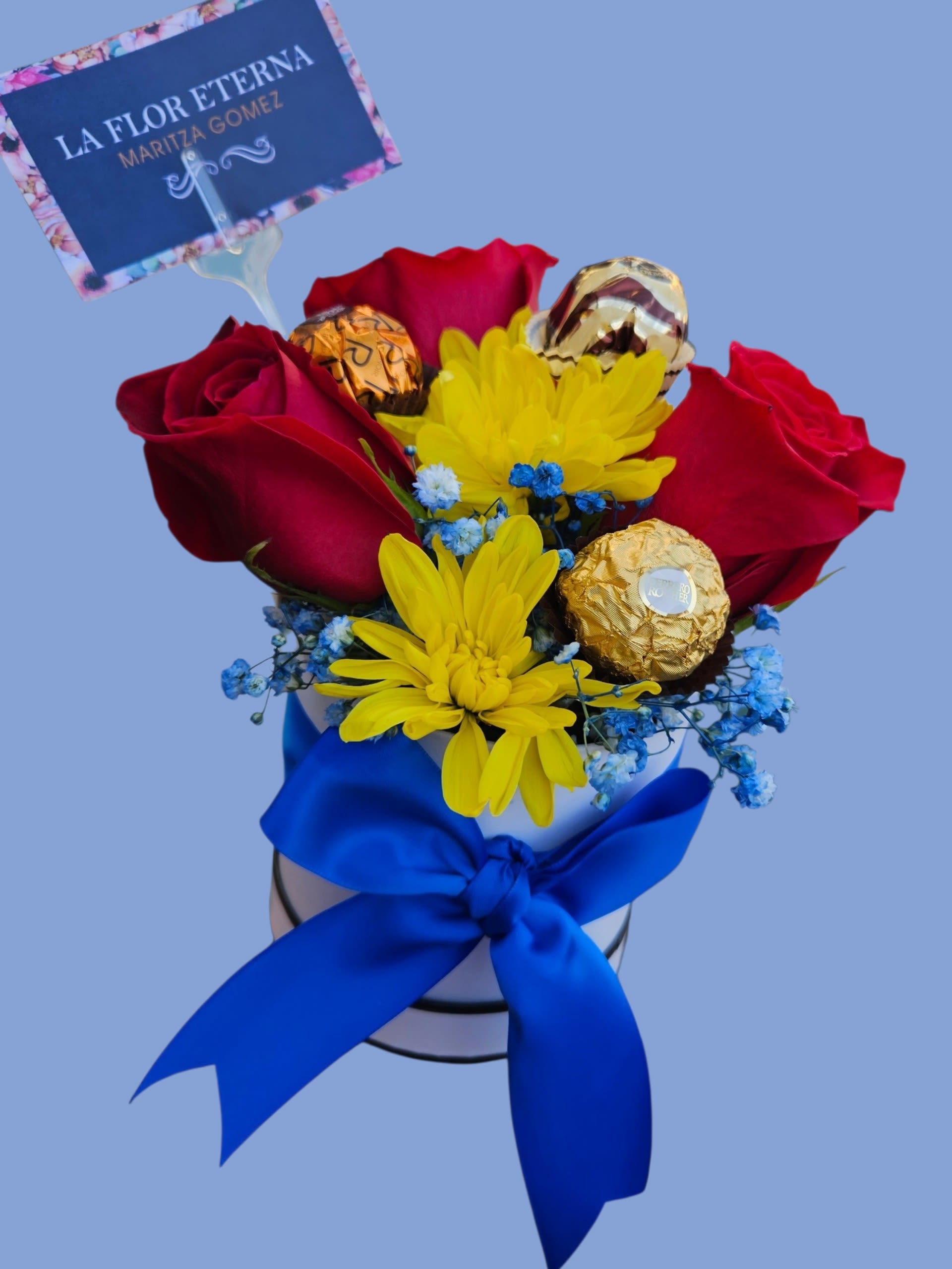 Forever Sweet - Deep red roses mingle with bright yellow daisies and tiny blue filler blooms in a compact round gift box-an arrangement that reads both romantic and celebratory, perfect as an anniversary bouquet or a cheerful birthday surprise. The mix of warm reds and sunny yellow with cool blue accents creates instant visual energy; the person you're thinking of will feel noticed and delighted the moment the lid lifts.  Tucked among the flowers are wrapped chocolate truffles and gold-wrapped chocolates, turning this into a floral-and-sweets gift set rather than a standard bouquet. Presented in a white hat-style box and finished with a large royal-blue satin ribbon, the package has a polished, gift-ready look that's easy to hand over or display. Dimensions follow the compact hat-box format; order with standard delivery options and include your personalized floral pick card to make the moment unmistakably theirs.
