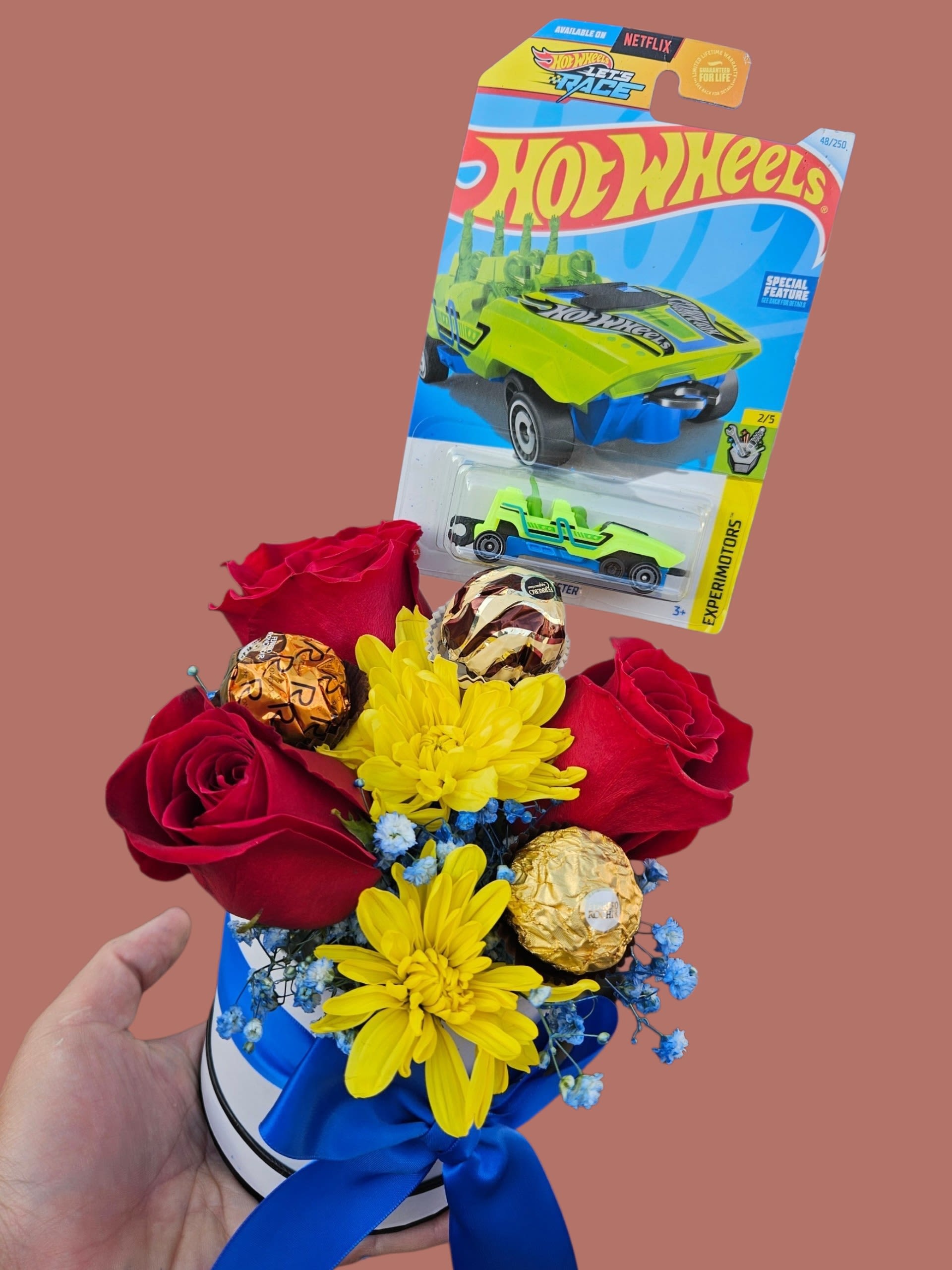 Amore Edible - A cheerful, playfully themed gift arrangement that feels like a mini celebration in a box-perfect as a birthday bouquet or a congratulations surprise. This flower arrangement combines deep red roses with bright yellow daisy-style chrysanthemums and tiny blue filler flowers to create a vivid, high-energy color contrast that reads fun and festive for a child or young-at-heart recipient.  Arranged compactly in a white round box and finished with a blue satin ribbon, the bouquet is accented with a Hot Wheels toy car package tucked above the blooms and several wrapped chocolate truffles nestled among the flowers. The mix of roses, chrysanthemums, daisies, and other seasonal filler gives a lively texture and cheerful palette (red, yellow, blue, white, mixed), while the toy and chocolates turn the piece into an instant gift set.  Give this to a child celebrating a birthday, a grad or milestone achiever who loves playful surprises, or anyone who'll appreciate a nostalgic, candy-and-toy twist on flowers. The compact size makes it easy to display on a table or desk; order with standard gift wrap and ribbon included for a ready-to-give presentation.