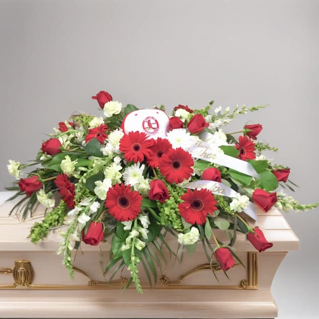Cardinals Fan Casket Spray - Hignight’s Cardinals Casket Spray: Honoring a St. Louis Cardinals Fan  In loving memory of a devoted St. Louis Cardinals fan, we present a breathtaking red and white casket spray of flowers.  As you bid farewell to a true fan, these vibrant blooms stand as a tribute to their unwavering love for the Cardinals. Each petal represents the countless moments of joy and excitement shared while cheering for their beloved team.  The rich red hues symbolize the passion that burned bright in their heart, while the pure white blossoms reflect the purity of their unwavering loyalty. Together, they create a striking and elegant display, befitting a true Cardinals enthusiast.  Our Cardinals Casket Spray serves as a reminder of the joy, unity, and everlasting spirit that the team brought to their life. Celebrating not only their love for the game, but also the memories and connections forged through it.  Please Note: Hat may vary depending on availability. Please call our shop if you would like to further customize this arrangement with your loved one’s personal sports memorabilia. This arrangement can easily be customized for any sports team of your choosing.