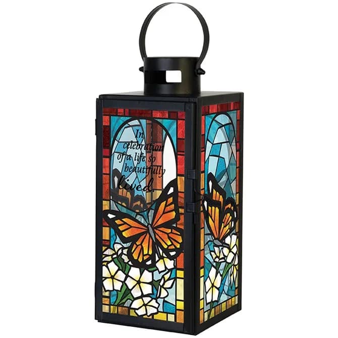 Celebration of a Life Stained Glass Lantern - This sympathy stained glass lantern features a durable black metal finish and elegant stained glass–style artwork, creating a meaningful memorial lantern illuminated by a flameless LED candle. Designed to cast a soft, comforting glow, it offers a beautiful way to honor and celebrate a life remembered.    This remembrance lantern can be displayed as a hanging lantern or used as a freestanding décor piece. Its weather-friendly construction makes it suitable for both indoor and outdoor use, including celebration of life services, memorial displays, gardens, patios, or as an accent in sympathy floral arrangements.    The lantern is inscribed with the heartfelt quote, “In celebration of a life so beautifully lived.”  A thoughtful sympathy gift, bereavement gift, or condolence keepsake, it pairs naturally with funeral flowers and remembrance décor.    Product Details:  Finish: Black metal  Artwork: Stained glass–style panels  Candle: Flameless LED  Display: Hanging or freestanding  Use: Indoor or outdoor  Power: 3 AAA batteries included       