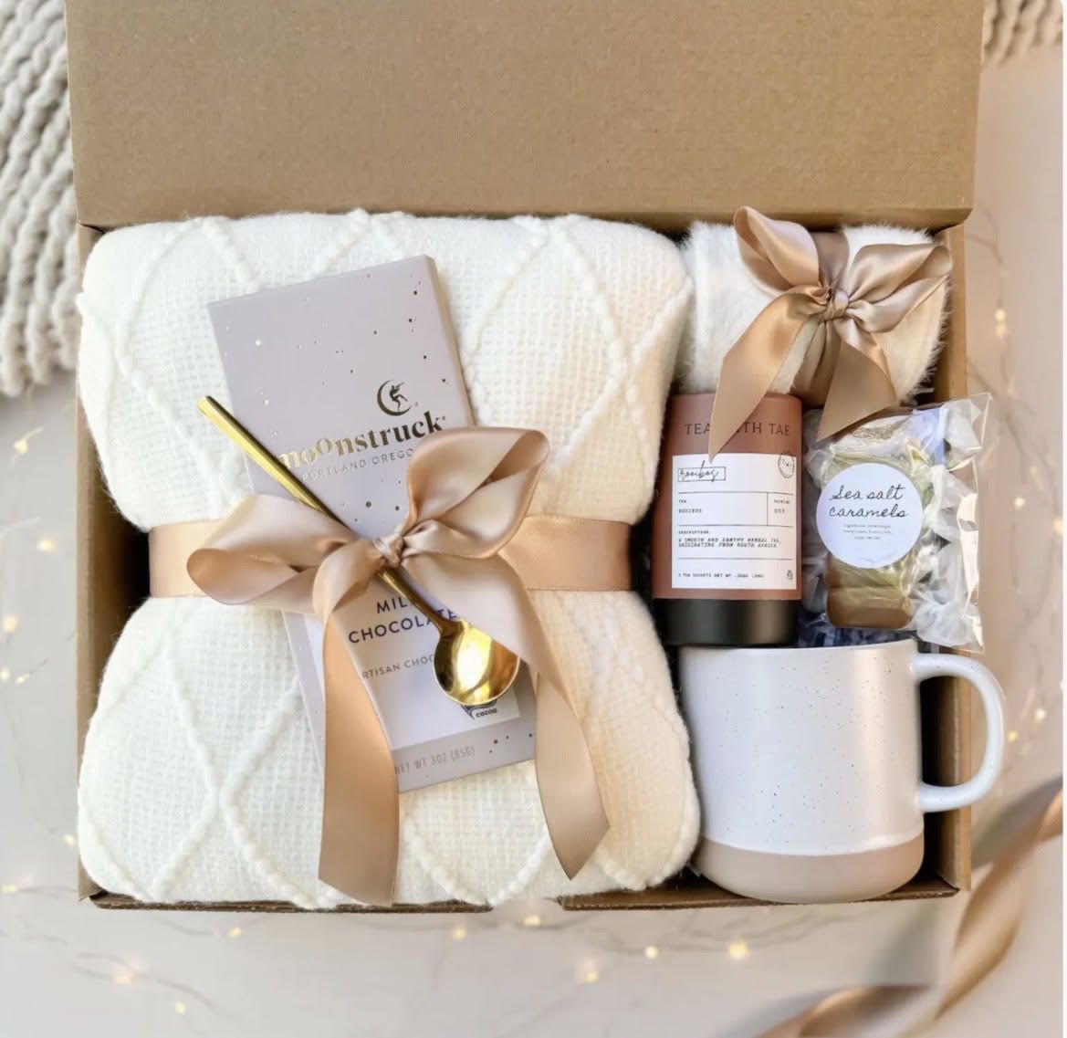 A cozy coffee break gift set - Wrapped in soft neutrals and gentle textures, this cozy coffee break gift set feels like a warm hug for the person you're thinking of. Designed as a gift set for birthdays, thank-you gestures, housewarmings, or get-well wishes, it combines comforting essentials and a subtle floral touch so the recipient feels genuinely cared for the moment they open it.  A shallow cardboard gift box holds a folded white knit blanket as the centerpiece, tied with a blush-peach satin ribbon that also secures a personalized gift card. Alongside the blanket sits a ceramic mug and gold spoon for slow sips, a tea tin and a chocolate bar for indulgent moments, plus a wrapped caramel for a sweet finish. The palette of white, pink, peach and soft black accents creates a calm, slightly luxurious look that reads as thoughtfully personal rather than overly formal.  This set is perfect for someone settling into a new home, recovering from illness, or simply deserving a quiet treat; it sends the message that you want them to pause and enjoy a peaceful moment. Sized to arrive ready to use, it requires no assembly-just tuck the blanket around someone you care about and let the small comforts do the rest.