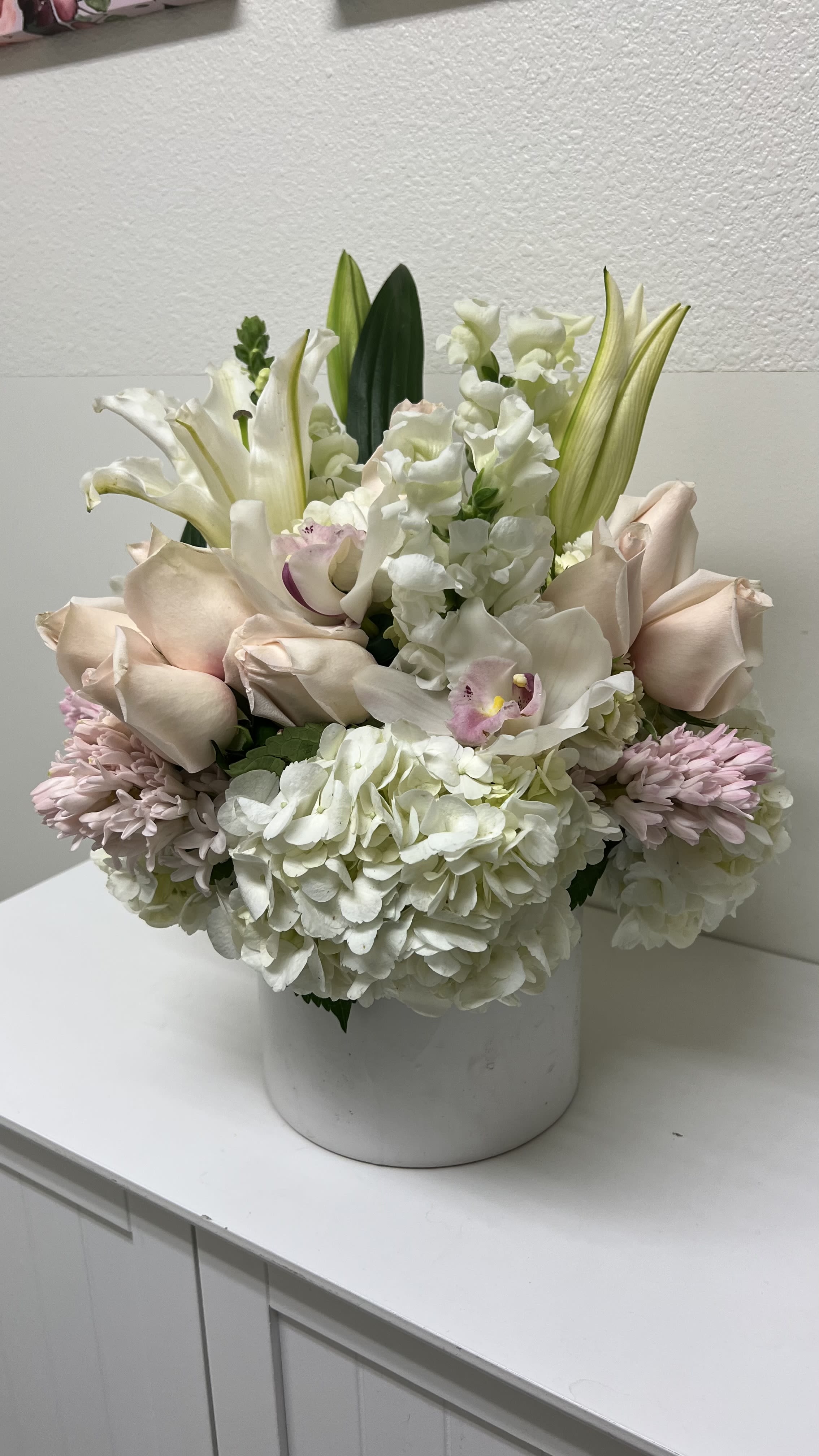 Angels in Bloom - Soft, airy white hydrangeas and blush roses form the heart of this sympathy arrangement, offering a gentle, comforting presence for memorials and moments of quiet reflection. Designed as a sympathy arrangement, the composition pairs white lilies and upright snapdragon spikes to give dignified height while pale pink chrysanthemums and subtle peach tones soften the overall feeling - a graceful way to express condolences and support.  Arranged in a rounded white ceramic vase, the bouquet is layered and full: the hydrangea mass anchors the base, blush roses and orchids add romantic warmth, and tall lily buds rise above for structure and serenity. The palette of white, pink, green, and peach reads refined and calming, suitable for a funeral service, visitation, or to send to the grieving person at home. This is the kind of arrangement that helps the recipient feel seen and gently comforted during a difficult time.