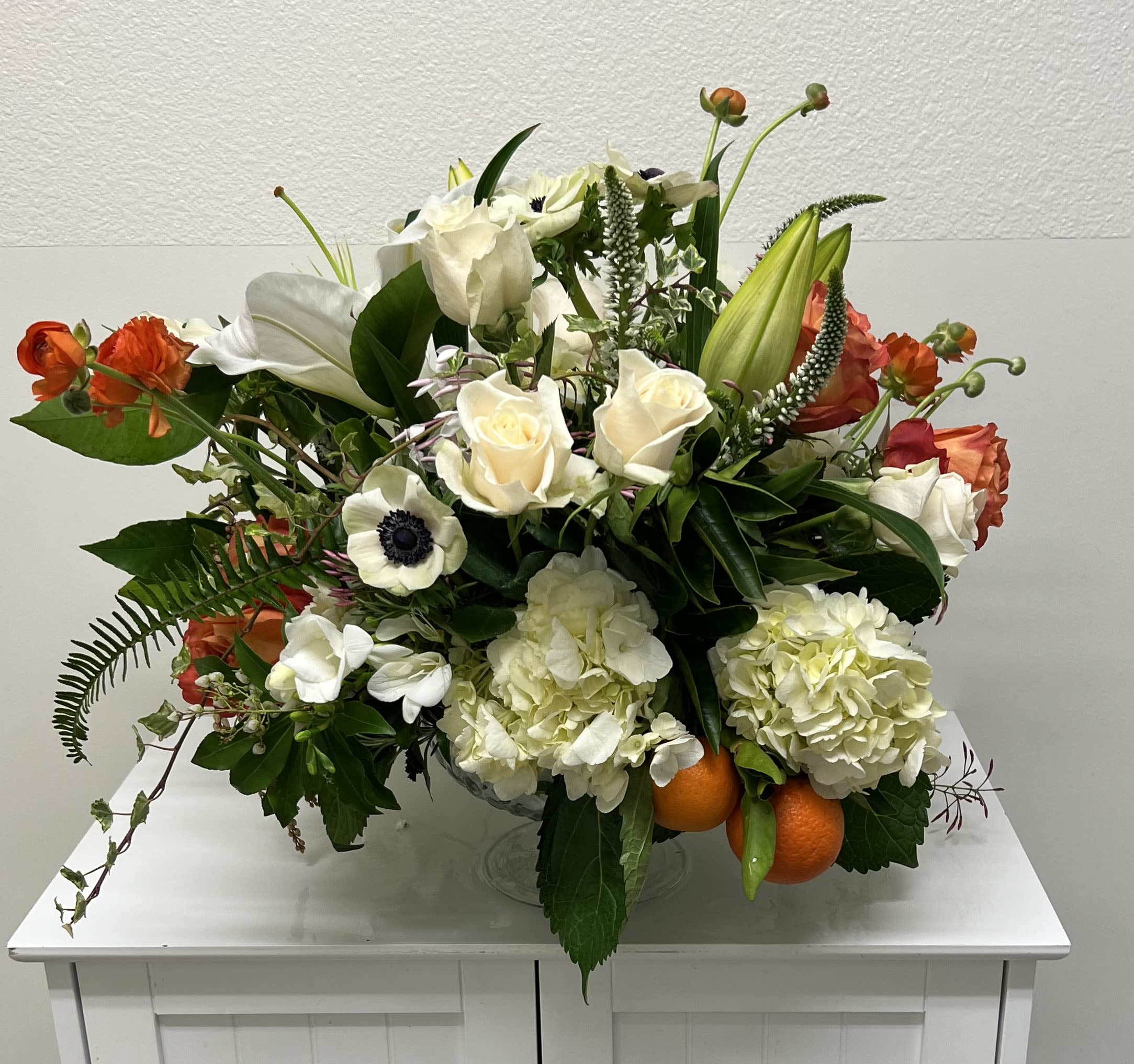 Eternal Life - A serene palette of creamy whites punctuated by warm orange and soft pink gives this sympathy flower arrangement a comforting, hopeful presence. As a sympathy arrangement designed to honor a life and offer solace, the low, lush composition reads as both elegant and approachable - the kind of display that helps the person you're thinking of feel remembered and supported during a difficult time.  Built full and layered in a glass compote vase, the design mixes white roses, white lilies, pale hydrangeas, tulip buds, and a white anemone with a dark center, with orange accent blooms and blush touches for gentle warmth. Dark glossy foliage, trailing ivy, and a few upright buds add height, movement, and textural contrast across the broad base. The result is a substantial, formal centerpiece with an airy, luminous quality created by the whites and creams against the vivid orange accents.  Appropriate for a memorial table, funeral service, or to send to a grieving household, this arrangement conveys sympathy and quiet celebration of a life. The low compote makes it easy to place on a viewing table or dining surface; standard care is to keep the vase filled with fresh water and away from direct heat to maintain presentation for several days.