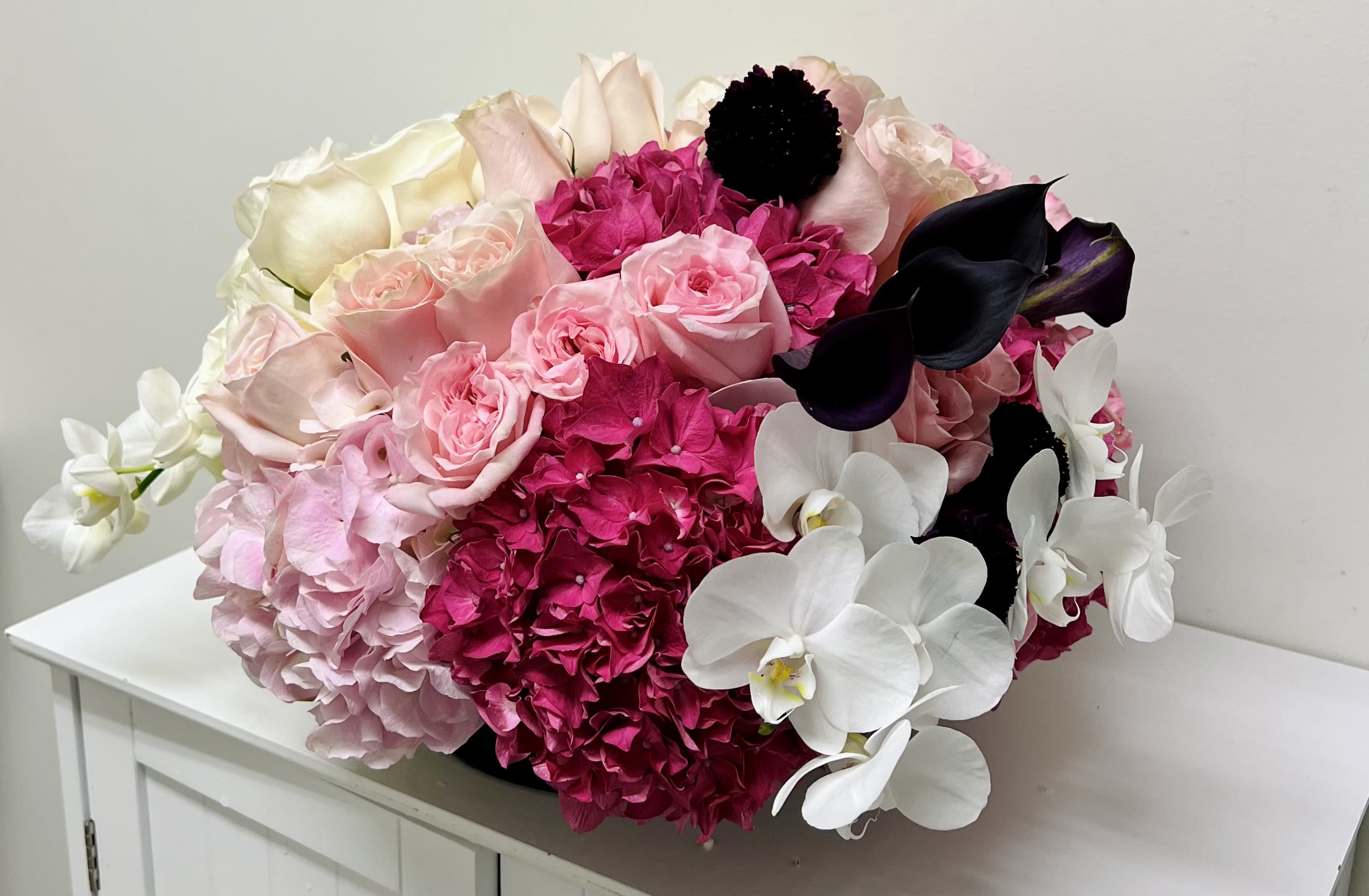 Everlasting  - A lush, rounded sympathy arrangement that speaks of gentle remembrance and quiet comfort, centered on soft pink and white blooms with dramatic deep magenta accents. This sympathy flower arrangement combines roses, hydrangeas, and white Phalaenopsis orchids to create a tender, elegant expression for a funeral service or to send to a grieving family-the kind of bouquet that lets the person you're thinking of feel honored and held during a difficult time.  Large hydrangea heads and layered pink roses form a full, cushion-like texture while crisp white orchids lend a refined, peaceful finish. Deep purple-black calla lilies and magenta highlights provide thoughtful contrast, giving the piece a polished, upscale look without feeling fussy. The design is compact and substantial rather than tall or airy, making it appropriate for a mantel, bedside table, or small altar space.  Intended as a sympathy gift, this arrangement conveys sympathy and respect while remaining versatile enough for memorial services or to send privately to a grieving home. It arrives ready to display in a low-profile style-full, cushioned, and arranged for immediate impact-so the recipient receives a composed, comforting statement at a time they need it most.