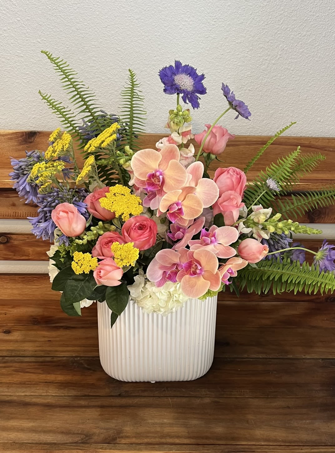Pure Pink - Soft peach and pink orchids paired with gentle pink roses and cloudlike white hydrangea create a serene, comforting sympathy arrangement that reads both tender and hopeful. Designed for funerals or to send condolences, this floral tribute uses a garden-like, slightly tropical mix of blooms to offer warmth without overwhelming - the kind of bouquet that helps the person you're thinking of feel remembered and supported.  The arrangement is composed of peach and pink orchids, pink roses, purple accent blooms, yellow filler flowers, snapdragons and white hydrangea, artfully arranged with tall fern fronds and airy stems for a full, asymmetrical silhouette. It arrives in a white ribbed ceramic vase that adds quiet texture and keeps the presentation restful and refined. The palette of soft pastels with brighter accents lends a gentle, uplifting note appropriate for springtime memorials or anytime you wish to express sympathy with grace.