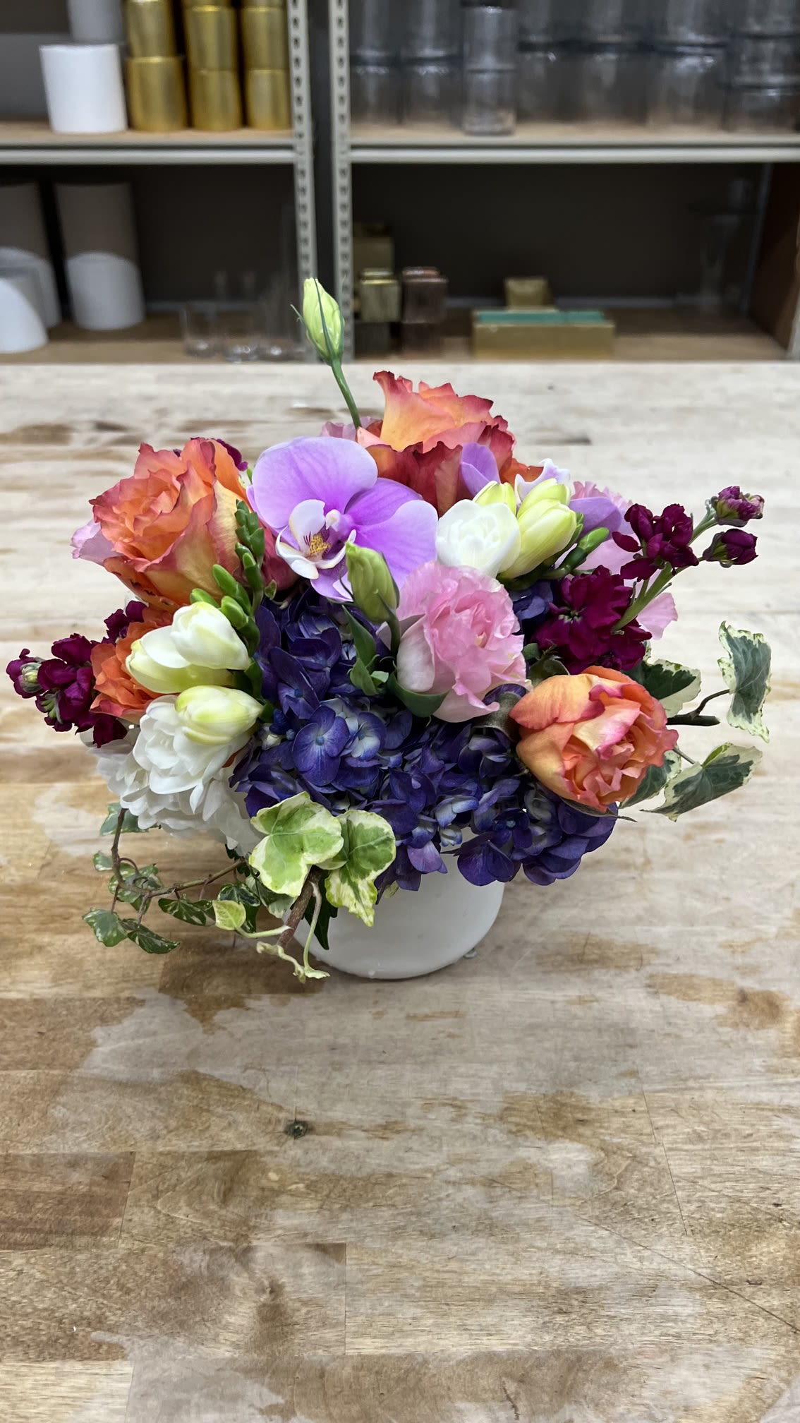 Colorful times - A bright lavender Phalaenopsis orchid anchored in the center gives this compact floral arrangement an elegant focal point that immediately delights the person you're celebrating. Designed as a birthday or "just because" gift, the rounded bouquet also reads beautifully for anniversaries, thank-you gestures, springtime brightening, or Mother's Day - it's the kind of arrangement that makes someone feel genuinely seen and cheered. The mix of purple, pink, orange, white and pale yellow colors creates a lively, celebratory impression without feeling fussy.  Closely clustered hydrangeas, orange roses, soft pink blooms, freesia and lisianthus form textured layers around the orchid, with a few arching orchid stems and buds adding graceful movement. Presented in a clean white ceramic vase, the design is compact and polished - easy to display on a desk, entry table, or bedside. The saturated, mixed-color palette reads as everyday festive yet refined, a versatile choice when you want to send warmth, thanks, or congratulations.  Sized as a small to medium tabletop arrangement, it's ready to gift with minimal fuss; to keep blooms fresh, place in a cool spot away from direct heat and change the water every 2-3 days. Whether marking a spring birthday, saying thank you, celebrating an anniversary, or surprising Mom "just because," this arrangement delivers an uplifting, memorable moment for the recipient.