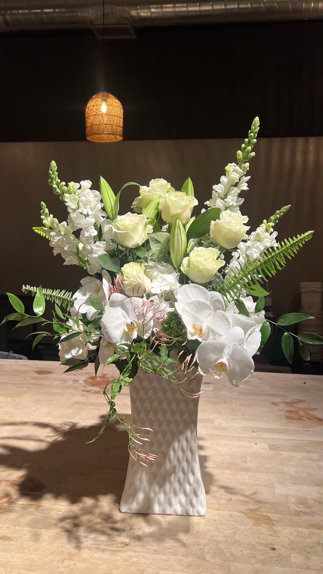 White Orchid Dreams - Creamy white phalaenopsis orchids and velvety white roses create a quietly elegant centerpiece that reads as both modern and romantic - perfect as an anniversary bouquet, wedding display, or sympathy arrangement. White orchids bring a sleek, sculptural presence while tall white snapdragon spikes add vertical movement; the overall effect is serene and uplifting for the person you're honoring.  This flower arrangement pairs those whites with glossy green foliage and delicate pink-tipped trailing stems that peek near the base, giving a soft hint of color and gentle motion. It arrives arranged in a tall textured white ceramic vase that reinforces the refined, airy composition and makes a statement on an entry table, altar, or dining room centerpiece.  Choose this for moments when you want someone to feel truly seen and comforted - a tasteful way to mark a milestone or offer sympathy. The tall ceramic vase makes placement easy; change the water every few days and trim stems as needed to keep blooms fresh longer.