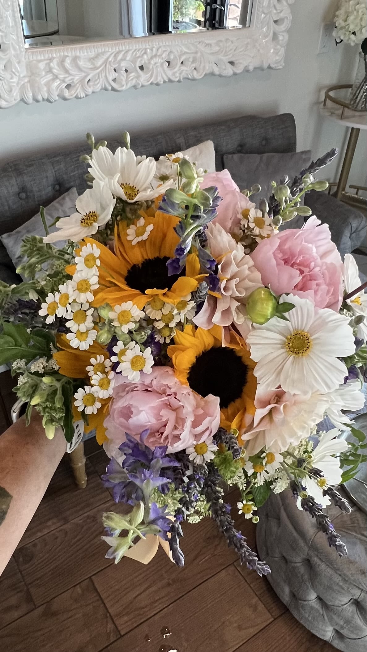 Sunflower Handheld  - Big golden sunflowers steal the show in this mixed-color flower arrangement, paired with soft pink peonies and clusters of small white daisy-like blooms for a sunny, garden-fresh look perfect for birthdays, thank-you gestures, or a romantic everyday bouquet. The combination of bright yellow, blush pink, white and subtle purple accents reads cheerful and slightly rustic, the kind of arrangement that makes the person you're thinking of feel genuinely seen and uplifted.  Flowers are arranged in a hand-tied style that's both full and airy-large sunflower heads give bold focal points while the peonies add plush texture and the daisies bring light, delicate detail. The dense-but-loose composition creates movement and a natural, just-picked-from-the-garden feel that works well for spring and summer gifting. Sized to be a standout on a table or counter, this bouquet arrives ready to display; change the water every couple of days and trim stems to help the blooms last longer.