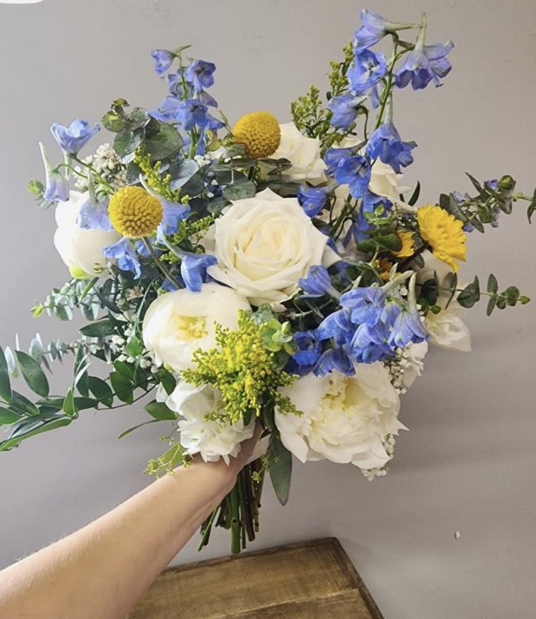 Blue Dream Handheld - Cool blue delphinium spikes and large white roses create a dreamy, romantic hand-tied bouquet perfect for a wedding bouquet or a celebratory gift. The Blue Dream Handheld reads fresh and airy - the tall blue accents lift the composition while pops of sunny yellow pom chrysanthemums bring cheerful contrast so the person you're thinking of feels both cherished and enlivened.  This arrangement pairs peony-like white blooms with classic roses, delicate delphinium, small yellow pom mums, and loose eucalyptus and mixed greenery for movement and texture. The bouquet has a relaxed, hand-tied shape that feels natural to hold and photograph; colors are white, blue, yellow, and green for a cool, uplifting palette. Ideal for brides, anniversaries, or to say "congratulations," it arrives ready to present-ask about size options or same-day delivery to ensure it fits the moment.