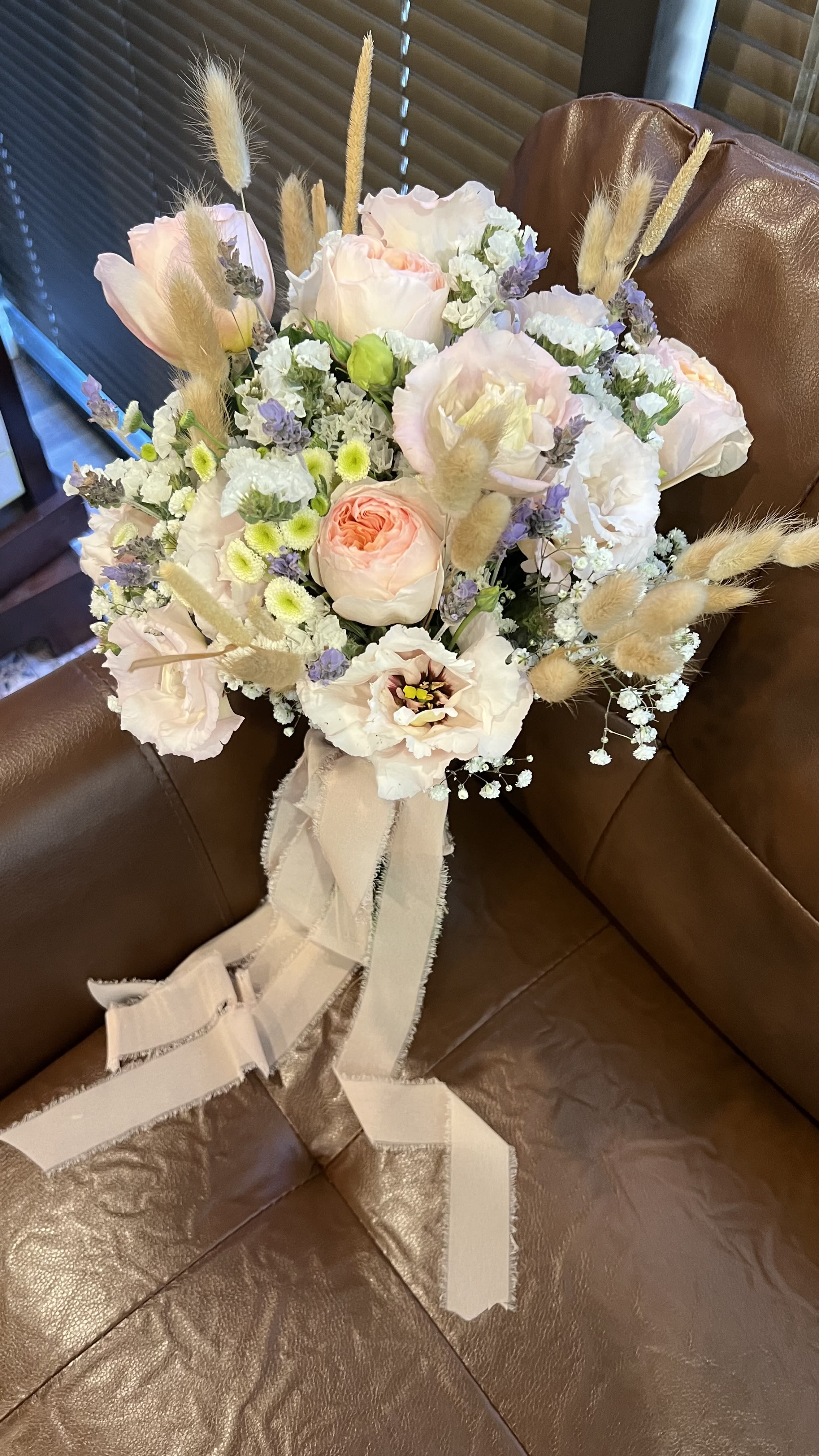 Country Handheld - Soft blush and ivory roses mingle with white lisianthus and pale chrysanthemum sprays in this Country Handheld bouquet, a romantic flower arrangement perfect for birthdays, anniversaries, or a thoughtful "thinking of you" gift. The gentle palette of pink, white, purple and a touch of yellow creates a delicate, airy impression that will make the person you're sending it to feel quietly celebrated and seen.  The loose hand-tied shape features airy lavender filler and dried bunny tail grass for a modern, textured accent that catches the eye without overpowering the blooms. Satin ribbon trails from the stems, lending an elegant, gift-ready finish. This bouquet feels light and refined-great for someone who prefers understated, natural charm over formal arrangements.  Designed to read as a refined everyday bouquet or a soft statement for special moments, Country Handheld is ideal for mothers, friends, or that special someone who loves a cottage‑garden look. Keep stems trimmed and fresh water available; the mixed blooms will hold well with regular water changes and a cool display away from direct sun.