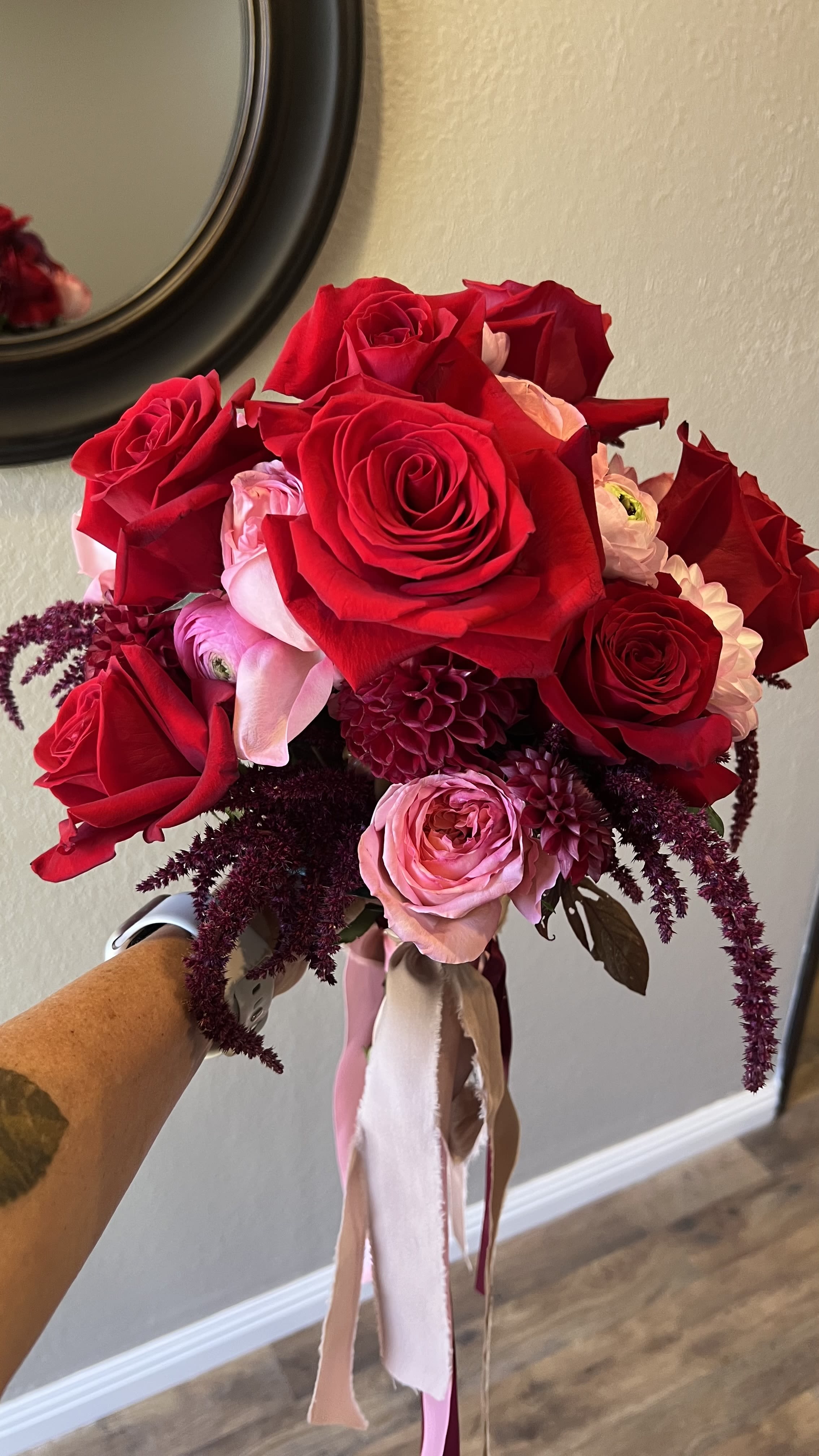 Passion - Velvety red roses take center stage in this romantic flower arrangement, paired with soft pink ranunculus and rich burgundy dahlias for a lush, celebratory bouquet perfect for an anniversary, Valentine's Day, or as wedding flowers. The mix of saturated reds, blush pinks, and touches of purple and white creates a deeply romantic color story that makes the person receiving it feel adored and truly seen.  This full, rounded design is hand-tied and finished with long pink satin and white ribbon tails for an elegant, dressed-up presentation. Expect layers of texture from the large roses, the delicate petal layers of ranunculus, and the bold form of dahlias-crafted to read as abundant and thoughtfully composed. Ideal for romantic gifting or marking a special moment, this arrangement arrives ready to display and delight.
