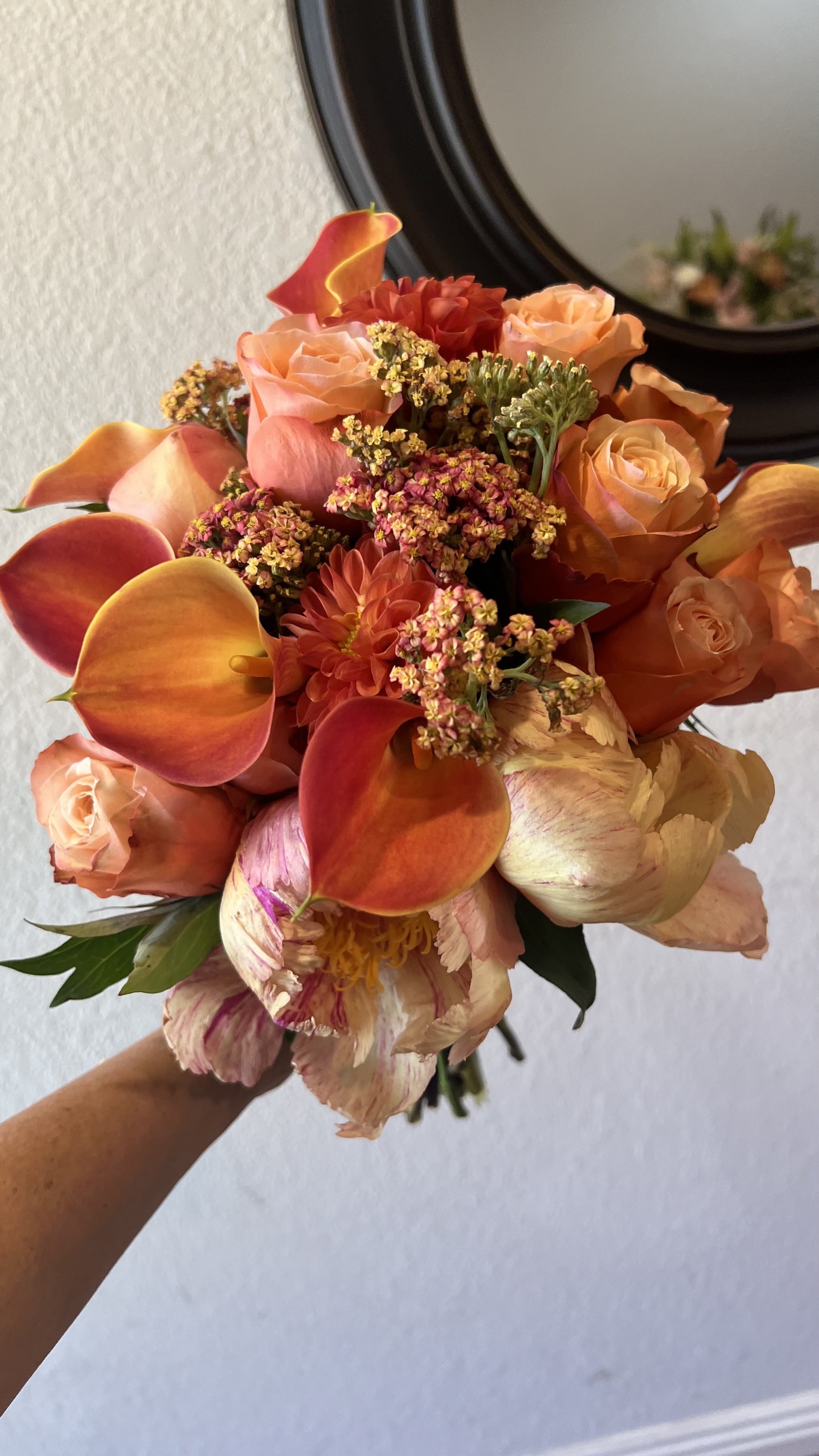 Rust - A lush rounded bouquet of warm coral, apricot and red tones that feels both romantic and celebratory - perfect as an anniversary roses-and-peonies arrangement or a wedding bouquet to make the recipient feel truly seen. The mix of peach roses and large pale peony blooms gives a soft, romantic center while vivid orange mini calla lilies and dahlias add energetic, sculptural accents; the overall impression is elegant but joyful, ideal for marking anniversaries, weddings, or special romantic moments.  This hand-tied composition is dense and rounded, with small clusters of tiny filler flowers woven through to create a slightly garden-like texture and natural movement. Colors range from soft peach and pale pink to rich rust and bright orange, so the bouquet reads warm and inviting from every angle. Sized to be a substantial, gift-ready arrangement, it arrives ready to display - keep stems trimmed and fresh water supplied to maintain the blooms for as long as possible.