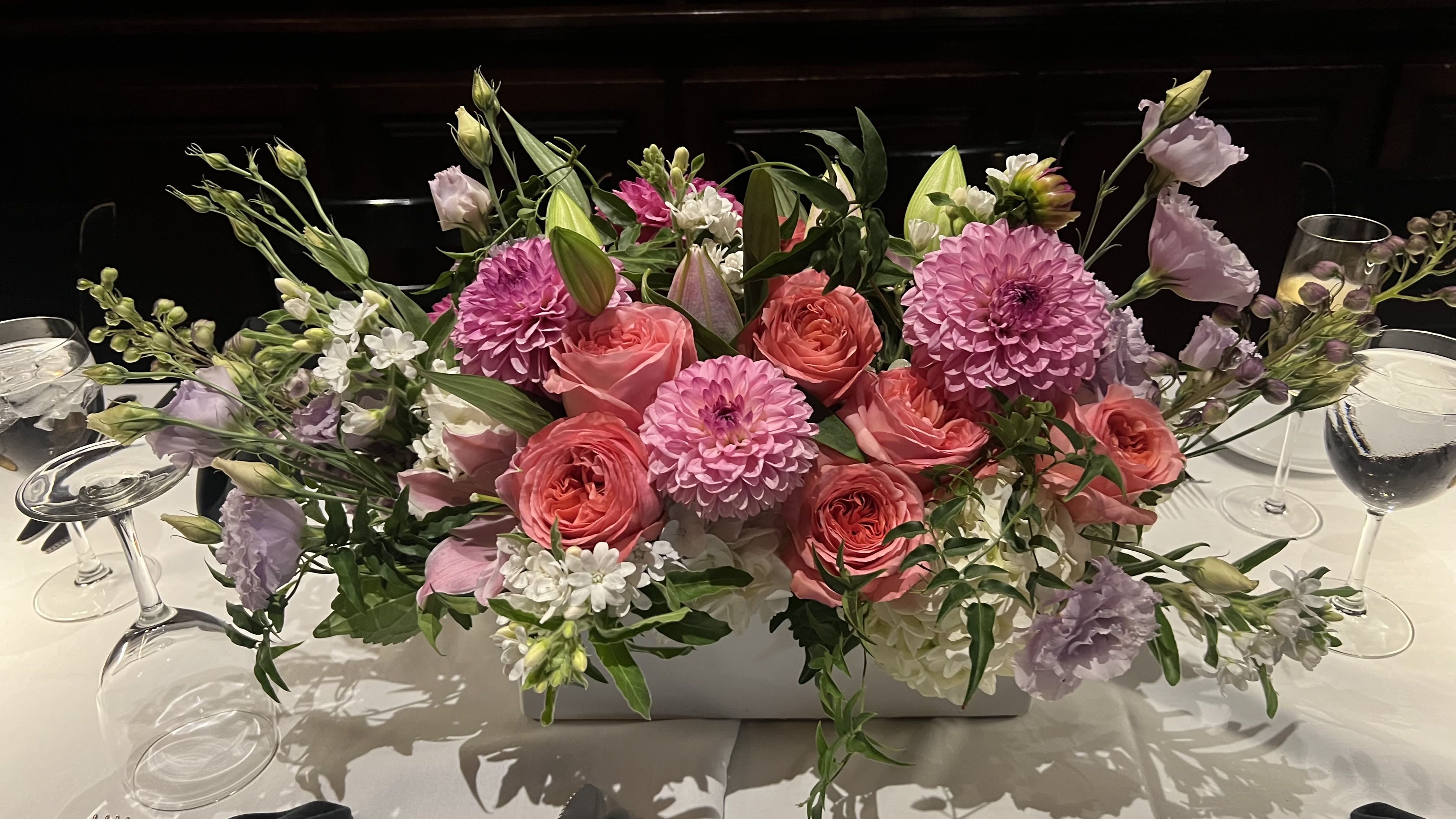 Perfect Pink - Soft pink roses and large pink dahlias create a lush, romantic centerpiece that reads like a spring garden across the table - ideal as a wedding centerpiece, anniversary arrangement, or for a celebratory dinner. The low, wide floral arrangement fills a white rectangular container and pairs beautifully with wine glasses and a crisp tablecloth, so the person you're honoring will feel genuinely celebrated without obstructing conversation.  Lavender lisianthus and small white filler blooms add airy texture and delicate contrast against the green foliage, while a few daisy-like blooms brighten the composition for a fresh, joyful note. The design spreads outward with blossoms and stems extending to both sides, creating an abundant, refined look that complements long tables or buffet displays. Its soft palette of pink, purple, white, and green keeps the focus on the recipient and the moment.  Perfect for spring gatherings, rehearsal dinners, or an intimate anniversary meal, this low-profile centerpiece makes the table feel thoughtfully dressed while keeping sightlines clear for guests. Available in the pictured white rectangular container; place on a protected surface and refresh water as needed to keep blooms looking their best during multi-hour events.
