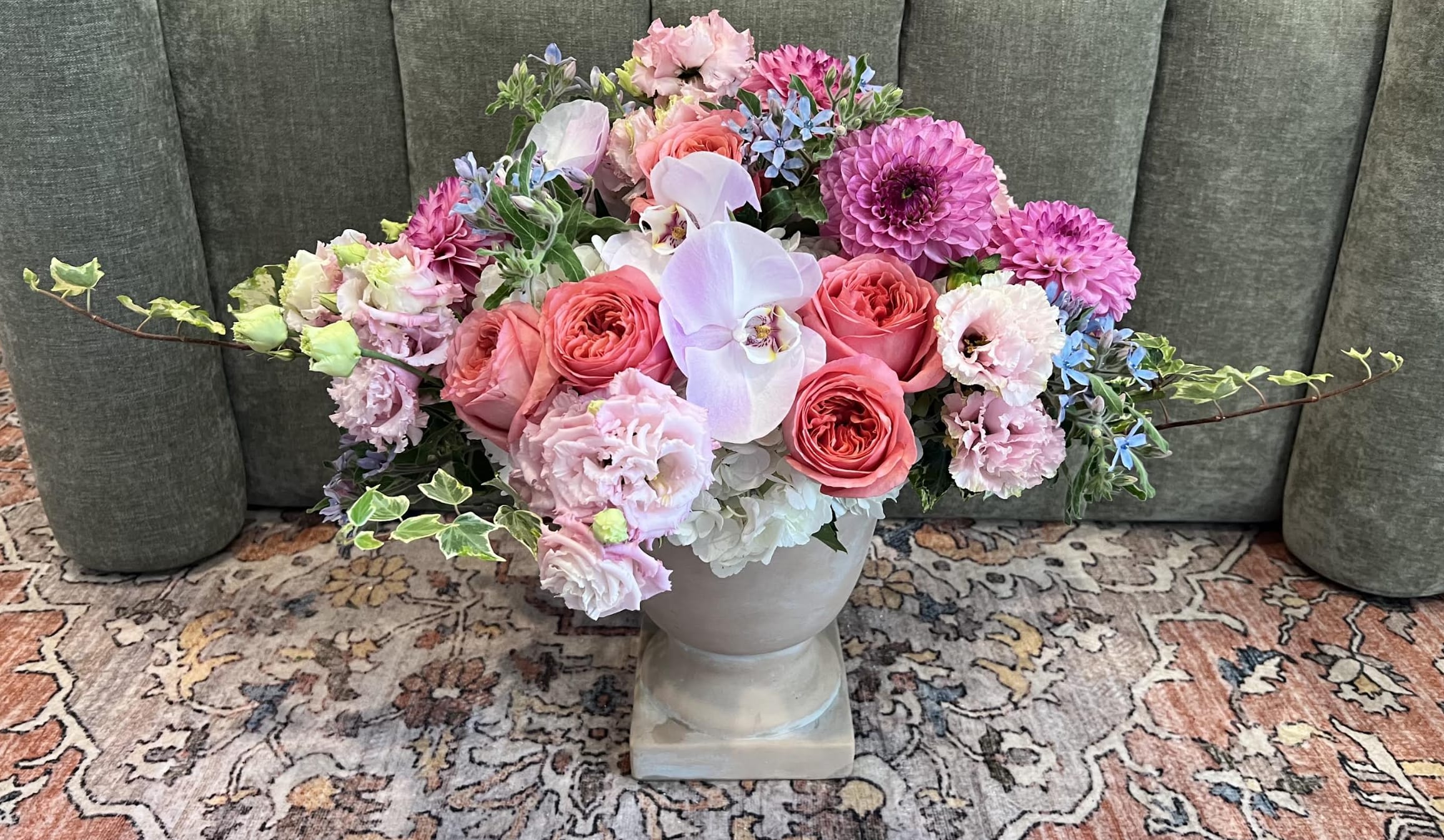 Paradise - A lush, garden-style centerpiece that reads like a breath of spring-this floral arrangement features coral-pink roses, bright magenta dahlias, and pale white orchids clustered with soft pink carnations and touches of blue filler blooms. Perfect as a wedding centerpiece, anniversary bouquet, or spring celebration arrangement, Paradise is designed to make the person you're honoring feel cherished and celebrated with its romantic, cheerful palette of pinks, whites, purples, and hints of blue.  Arranged low and wide in your choice of a pale gray ceramic vase or a clear glass vase, the composition is layered for texture: large coral roses and ruffled dahlias anchor the display, orchids add elegant airiness, and hydrangea-like massing plus small blue accents lend contrast. Trailing, variegated ivy-like stems and airy branching details give the piece a slightly whimsical, garden-fresh silhouette that looks abundant from every angle.  Ideal for a bridal table, anniversary dinner, or to brighten a special someone's day, Paradise blooms are arranged for immediate impact and easy care-kept in fresh water and away from direct heat they'll stay vibrant through the celebration. Choose the vase that best fits the setting and order with confidence to create a memorable, romantic floral moment.