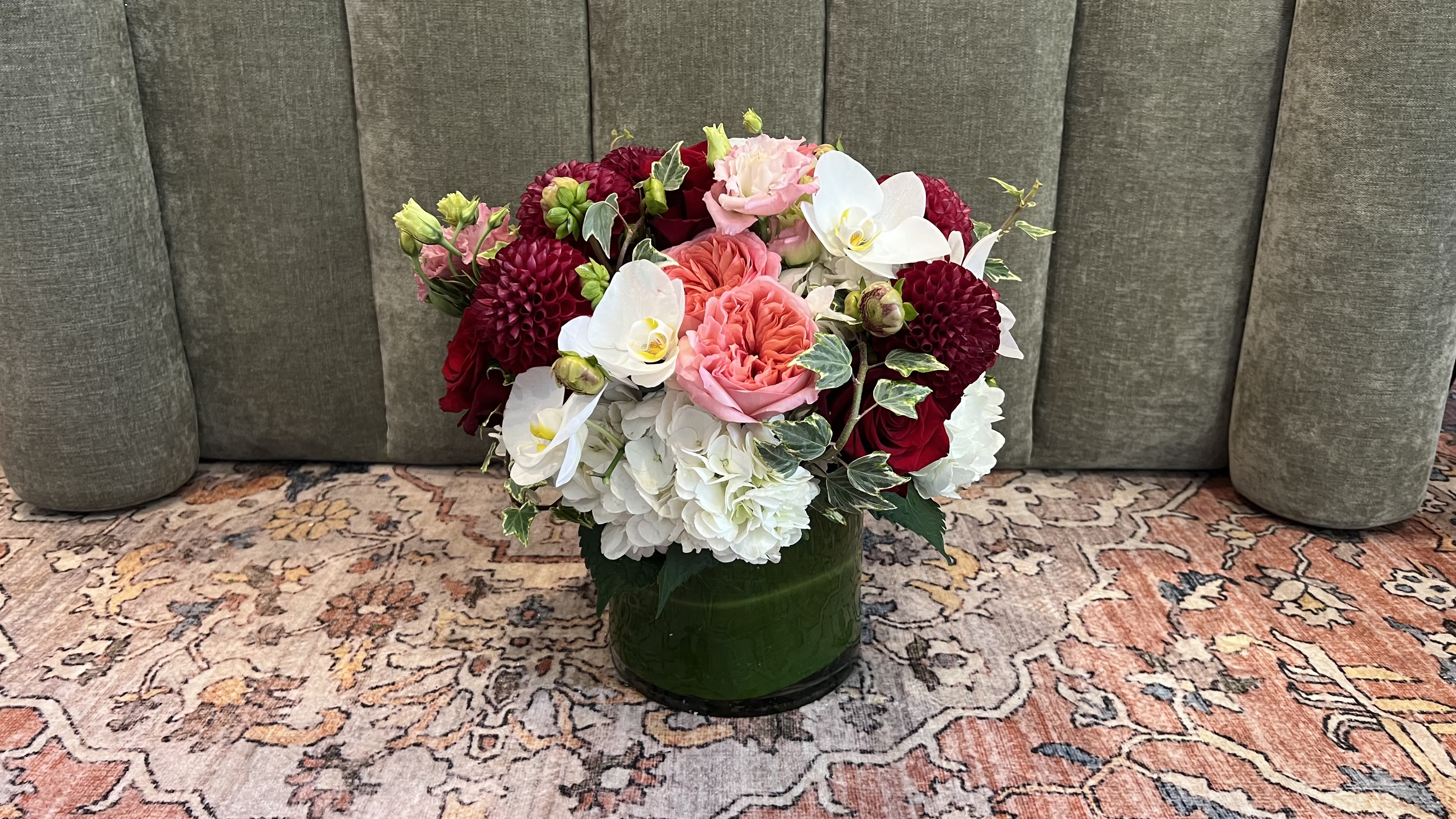 Ruby - A lush, jewel-toned rounded arrangement designed to make her feel truly celebrated-perfect as an anniversary bouquet, birthday gift, or just-because surprise. This flower arrangement blends romantic pink and coral roses with deep burgundy dahlia-like blooms and crisp white phalaenopsis orchids, giving the piece both softness and dramatic contrast. The compact, low profile reads polished and intimate, the kind of centerpiece that says "you are seen" when it arrives at her table.  Packed tightly for a full, textured look, Ruby combines white hydrangea and pale pink carnations to create a creamy base that lifts the darker blooms, while coral and blush roses thread warmth through the composition. Variegated ivy trails around the edges for graceful movement and a hint of greenery. The design is presented in a low, dark green glass vessel (also shown in low black container options), making it a perfect everyday centerpiece for mantels, dining tables, or bedside tables.  Ideal for birthdays, anniversaries, thank-you gestures, or spring celebrations, this arrangement is built to draw compliments and linger in memory. The compact, rounded silhouette keeps stems snug for longer-lasting freshness; change the water every two days and trim stems slightly if relocating to a new vase to extend enjoyment. Order with confidence for a meaningful, polished gift that matches romantic moments and cheerful occasions alike.