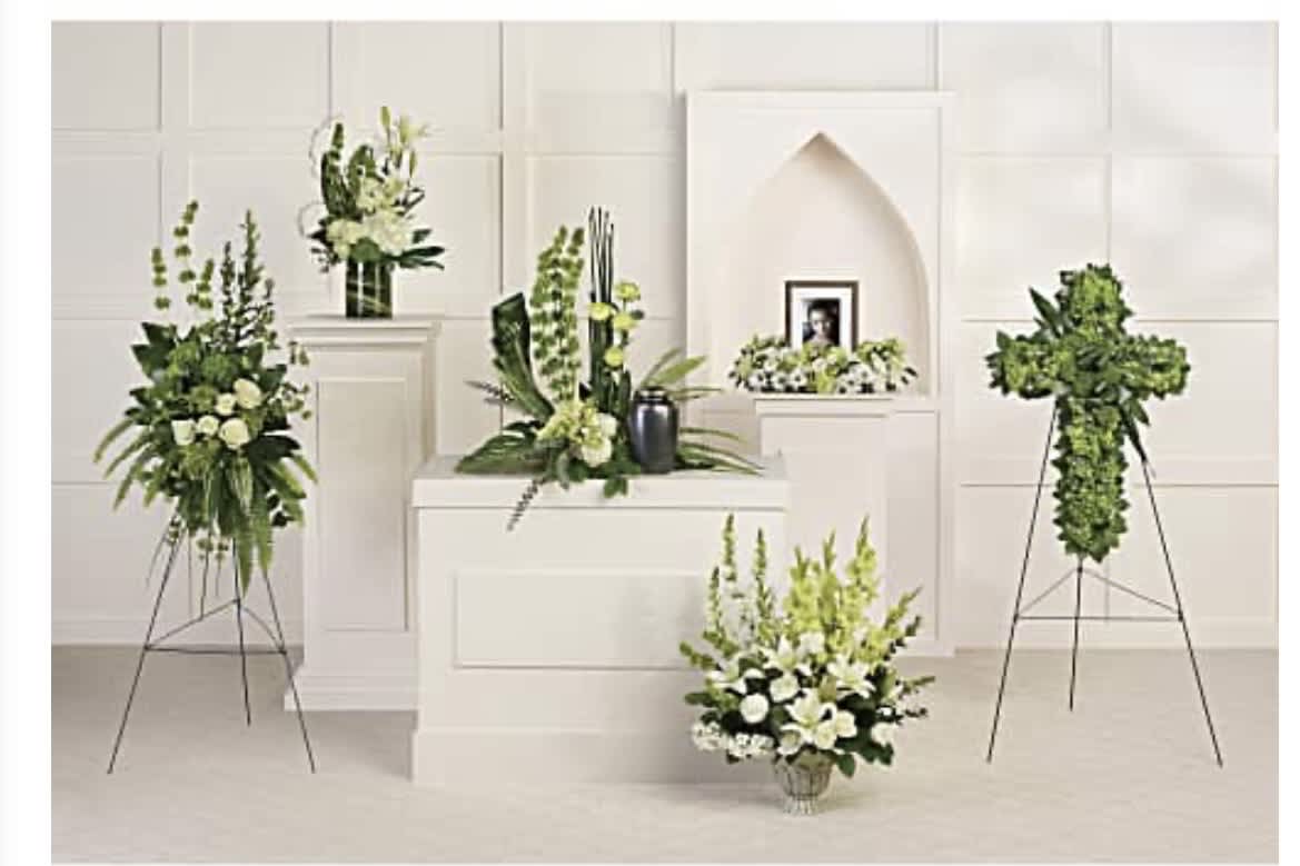 The Eternal Legacy Urn Package - A serene coordinated memorial set that honors a life with quiet dignity - perfect for a sympathy tribute or funeral service. This arrangement package pairs upright sprays and fuller low arrangements to create a balanced display around a black urn and framed photo, giving the person you're remembering a calm, formal presentation that feels respectful and composed.  White lilies, roses, chrysanthemums and daisies are arranged against abundant green foliage to emphasize purity and comfort; mixed-color accents are used sparingly to add gentle warmth without distracting from the memorial focus. The designs come presented on white display pedestals and metal easels with pedestal stands to suit chapel aisles or a home memorial table, creating a cohesive look across multiple pieces.  Choose this set when you want the family to feel supported and honored - it's the kind of tribute that helps guests pause, remember, and find solace. Sizes and specific piece counts are arranged to match the service setting; please contact us for delivery timing, personalization (including placement of the framed photo), or adjustments to fit the space.