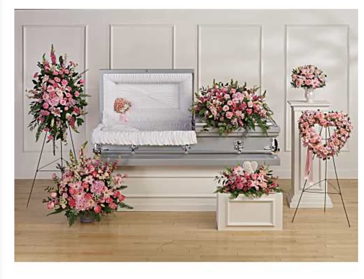 The Grace Collection Urn Package - Soft pink roses and lilies arranged with white carnations and alstroemeria create a serene, ceremonial presentation designed for a funeral service or memorial - a graceful funeral arrangement set that comforts and honors. The Grace Collection Urn Package frames the casket and service area with coordinated sympathy pieces so the person you're remembering is surrounded by gentle color and dignified formality.  This package includes a large casket spray plus matching standing sprays, wreath-style tributes, and urn arrangements presented on a white display pedestal and pedestal vase as shown, with options for standing easels. Blooms are densely layered for a lush, polished look: soft pinks balanced by crisp whites and rich green foliage to convey tenderness and respect. The mixed bouquets and traditional flower varieties (roses, lilies, carnations, alstroemeria) are arranged for lasting visual impact throughout the service.  Appropriate for a formal funeral or memorial ceremony, The Grace Collection offers a cohesive, respectful tribute that helps the family feel supported and honored. Please indicate delivery details, service time, and any personalization requests when ordering so we can coordinate placement on the casket, easels, or urn pedestal.