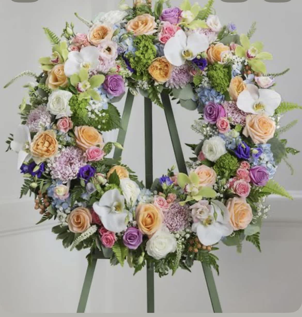 Summer Gardens - A lush circular wreath of pastel roses, Phalaenopsis orchids, hydrangeas, lilac and ranunculus creates an instant garden moment-perfect as a wedding floral display or a celebratory sympathy piece. This large standing flower arrangement announces itself with layered white orchids and velvety roses in soft pinks, peach and lavender, giving the recipient a feeling of gentle elegance and heartfelt thoughtfulness.  Built as a full, garden-style surface on a wreath stand, the design uses densely packed blooms and textured fillers to produce a refined, sculptural silhouette that reads beautifully from every angle. The palette blends white, pink, purple and touches of soft blue; the mix of round hydrangea clusters, ruffled ranunculus and delicate lilac adds depth and a soft, fragrant texture that complements the orchids' sleek petals.  Ideal for honoring a milestone, commemorating a life, or anchoring a wedding ceremony backdrop, this arrangement makes someone feel truly seen and celebrated. Because it's made to stand, it's sized for formal display-please indicate delivery placement and any dedication message when ordering so we can ensure the presentation matches the moment.