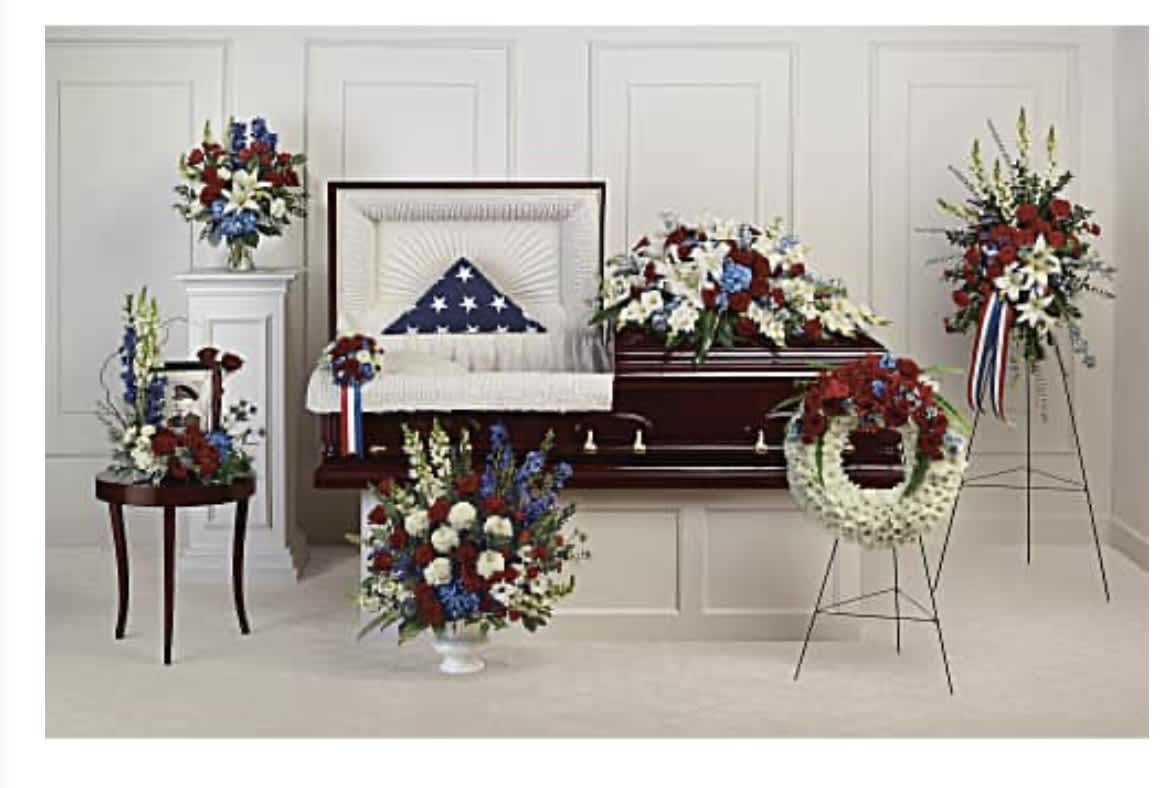 The Honor Urn Package - A striking red, white and blue funeral arrangement designed to honor a veteran or mark a patriotic remembrance, The Honor Urn Package creates a solemn, ceremonial atmosphere for a memorial service. This funeral set centers on a polished casket draped with an honor flag and finished with a large casket spray of red roses, white lilies and carnations with blue delphinium accents-an arrangement meant to convey respect and gratitude to the person you're remembering.  Coordinated matching pieces complete the display: standing easels with sprays, a white chrysanthemum wreath, a pedestal table arrangement, and a small side-table vase and framed display to accompany the urn or service materials. Lush greenery ties the group together, giving depth and texture while keeping the focus on the red, white and blue palette. Designed to fill a service space with dignity, these pieces are sized for full-ceremony presence and arranged by our florists for a balanced, reverent presentation; please call the shop for delivery timing, setup details, or to discuss personalization for military honors.