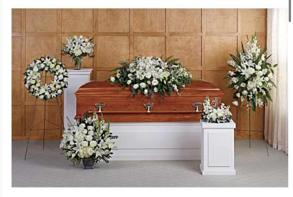 The Everlasting Tribute Urn Package - A serene, all-white tribute crafted for a funeral or memorial service, this flower arrangement package centers on timeless sympathy florals to honor a life with dignity. The Everlasting Tribute Urn Package is designed specifically for a chapel setting and for use with a wooden casket or urn, creating a cohesive, respectful display that helps the family and guests feel comforted and remembered.  White lilies, roses, carnations, chrysanthemums, and alstroemeria are arranged into a large casket spray, standing wreaths, and elegant pedestal pieces; deep green foliage shapes each composition and lends a formal, structured silhouette. The set includes matching standing easels and white pedestal stands to present the arrangements at full height around the polished wooden casket, plus a complementary vase or urn arrangement when requested. The overall color palette-pure whites with dark green accents-keeps the focus on quiet beauty and reverence.  Perfect for a traditional funeral, memorial service, or celebration of life, this package conveys solemn respect and heartfelt sympathy for the person you're honoring. For placement and sizing to suit your chapel or viewing room, please specify casket or urn preference and service details when ordering so we can prepare the arrangements and provide delivery and setup assistance.