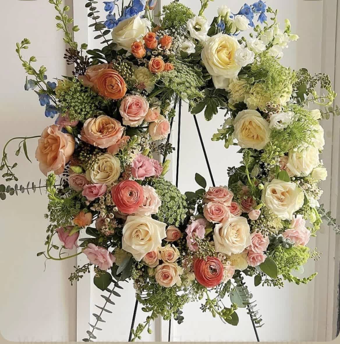 Spring Wreath - A lush, garden-style spring wreath that reads as both celebratory and elegantly romantic - perfect as a sympathy tribute, memorial display, or meaningful centerpiece for a springtime gathering. This flower arrangement wreath blends roses and ranunculus with daisies and airy fillers in a soft palette of white, pink, green, and touches of blue, giving the person you're honoring a gentle, uplifting presence.  The design is densely packed in a large circular form, with cream, blush, peach, coral and soft blue blooms layered for depth and texture; trailing greenery and delicate filler flowers add movement and a natural, hand-crafted look. Roses and ranunculus dominate the composition, supported by daisies and seasonal accents to create a full, abundant silhouette that reads beautifully from every angle.  Presented on a sturdy metal stand, this wreath is intended as a decorative display piece for services, memorial corners, or an elegant focal at a reception. It arrives ready to display; keep it in a cool spot out of direct sun to extend freshness, and contact us if you need help with delivery timing or stand placement for your event.