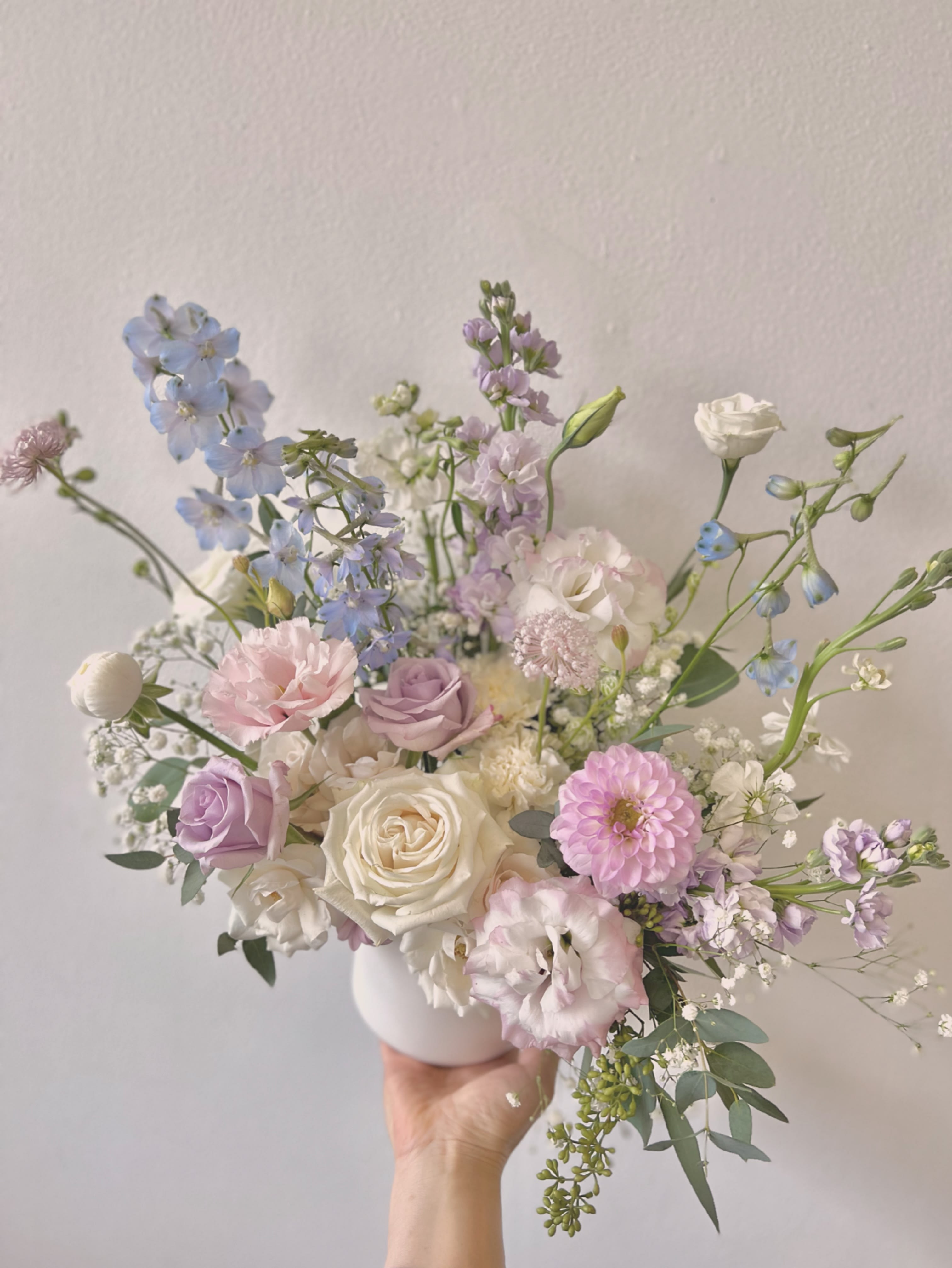 Pastel bouquet of roses, lisianthus, and blue delphinium in a white vase