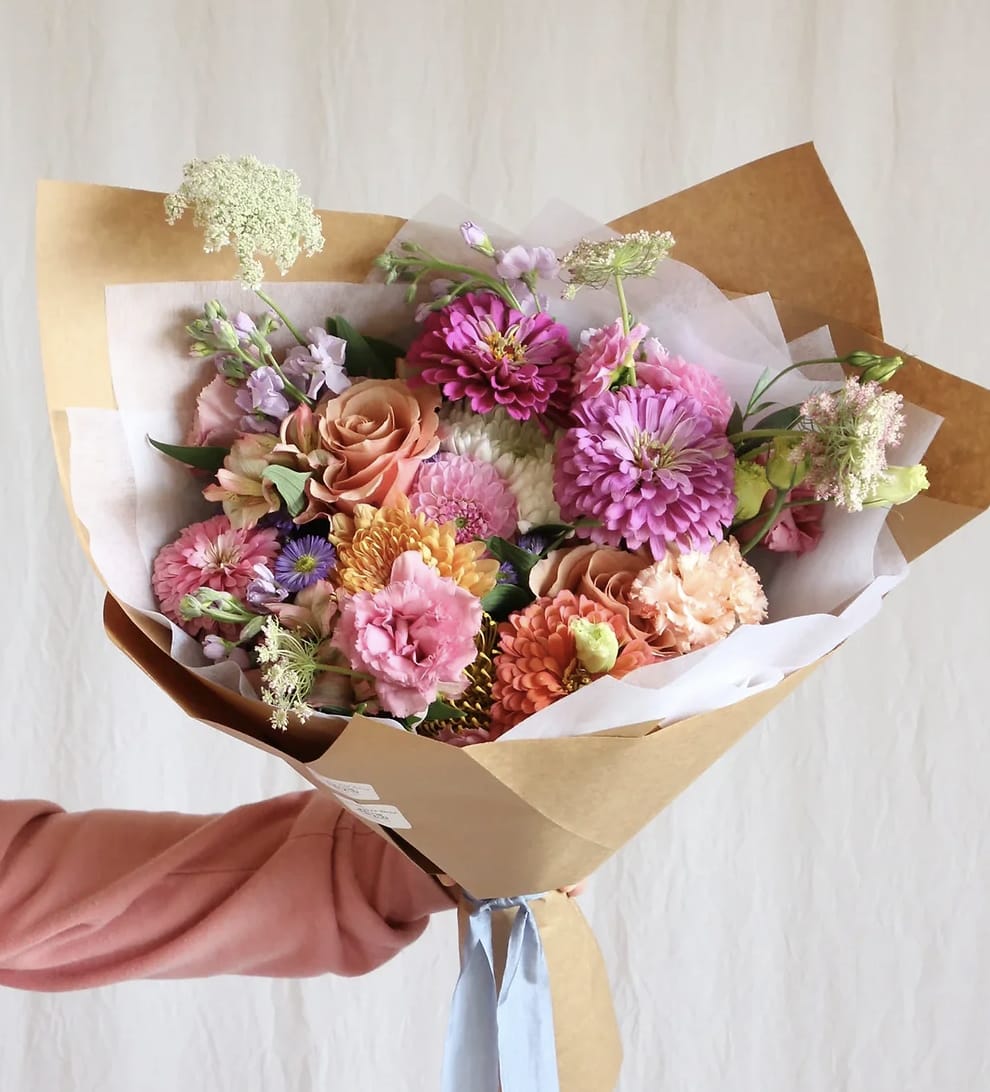Hand-tied bouquet of pink, peach, and purple flowers wrapped in brown paper