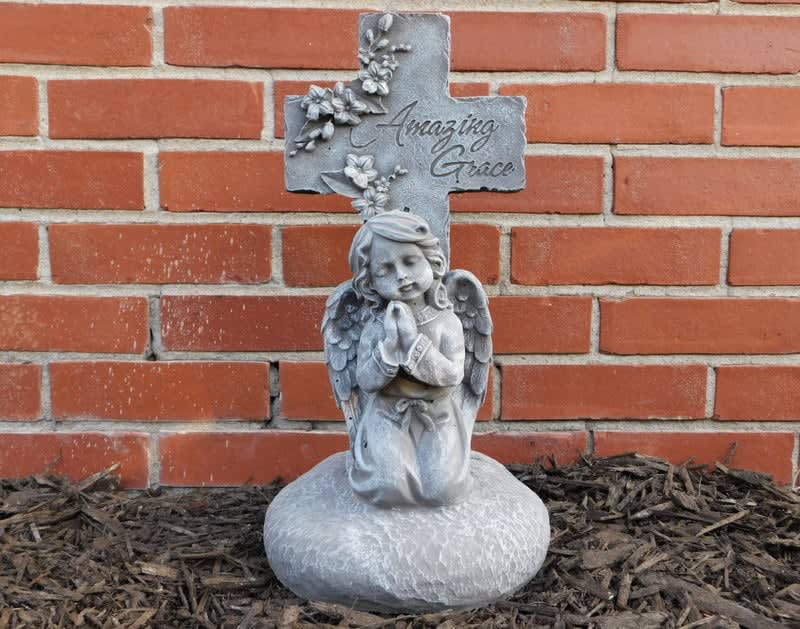 Gray angel statue kneeling before a cross with flowers and the words Amazing Grace