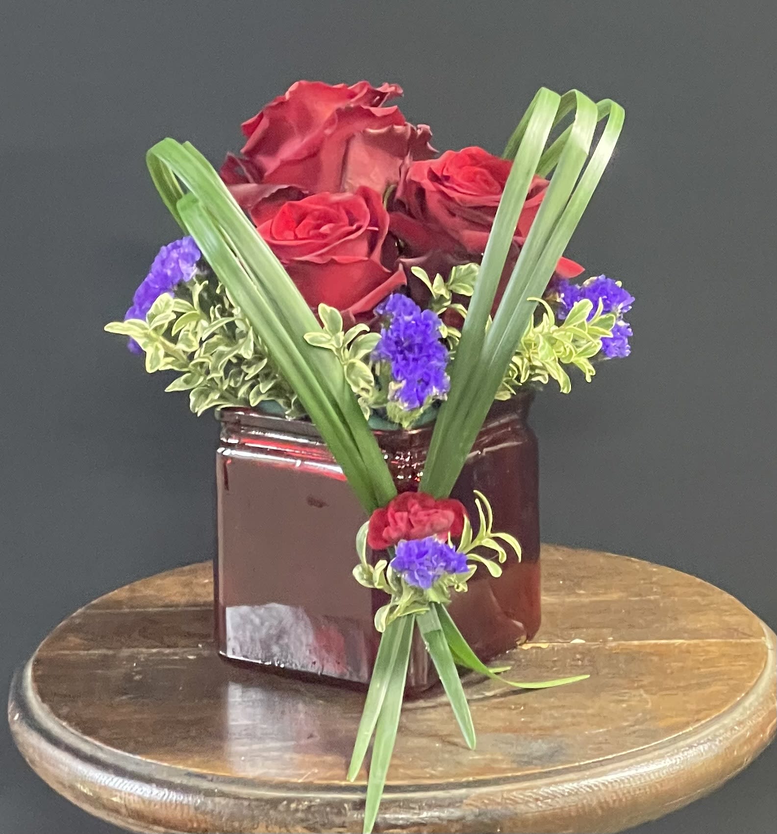 Red roses in a burgundy vase with purple accents and long green leaves