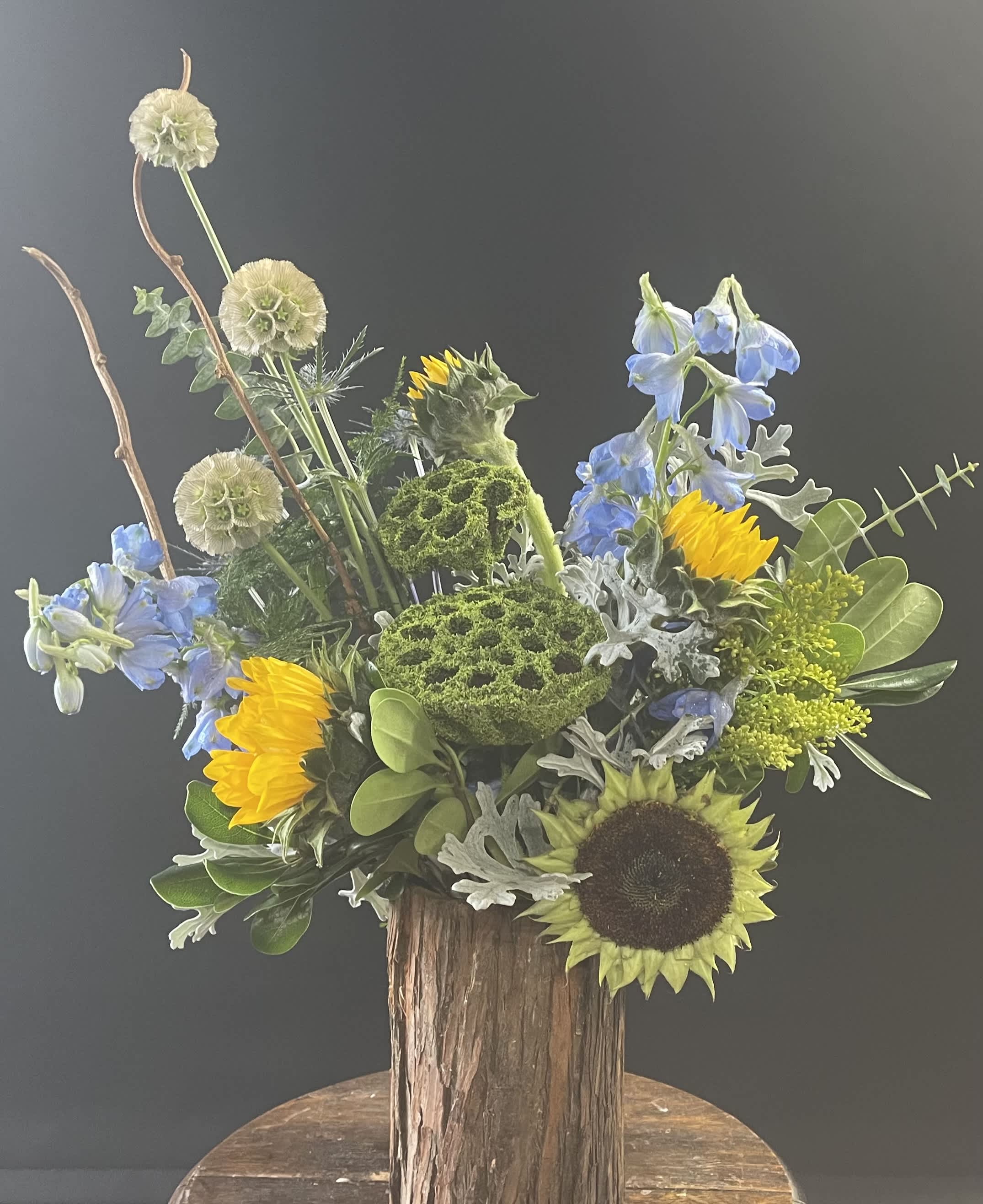 Mixed bouquet with sunflowers, blue flowers, and green textured blooms in a rustic wood vase