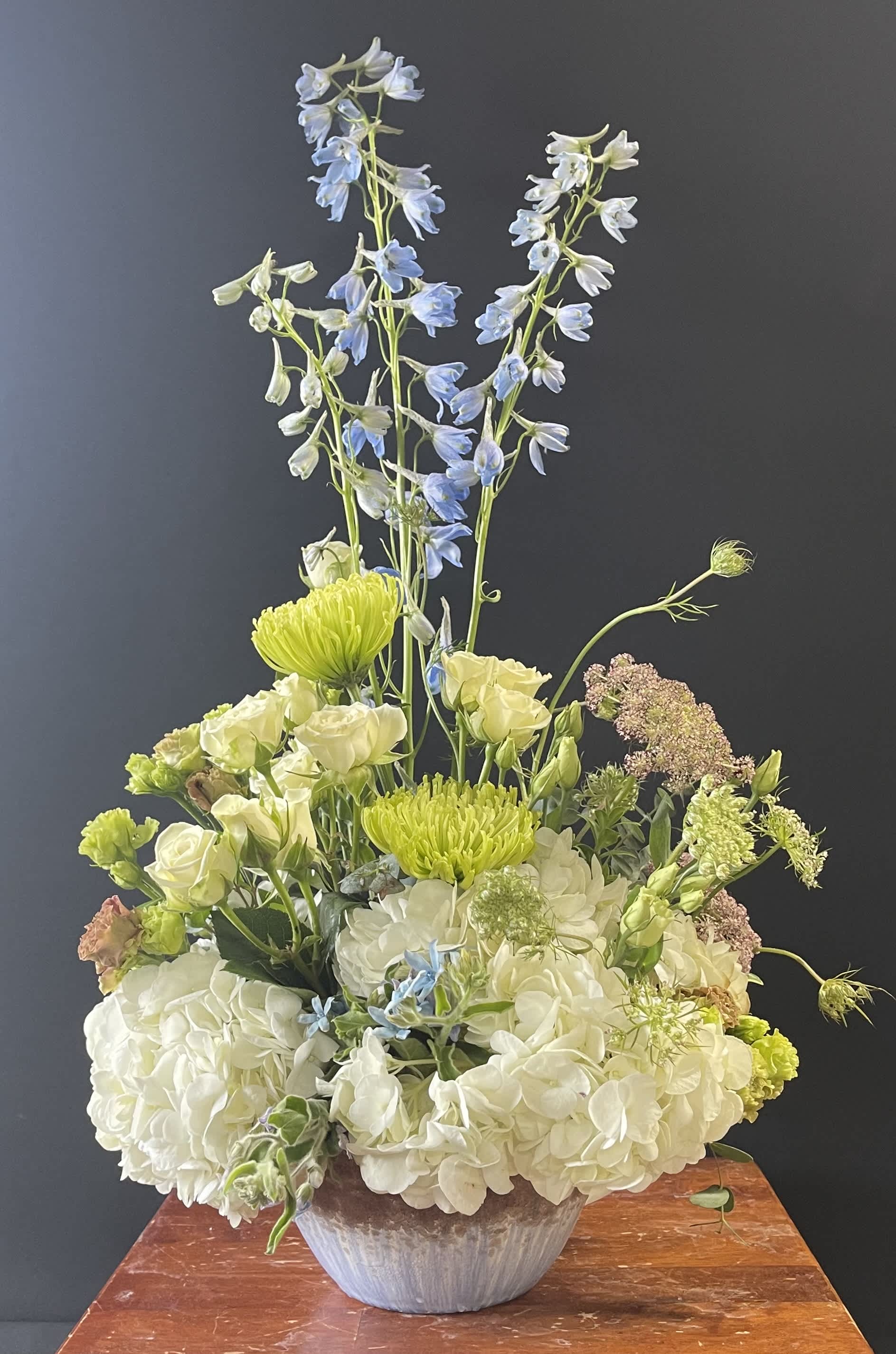 Tall floral arrangement with white hydrangeas, blue delphinium, and green blooms in a vase