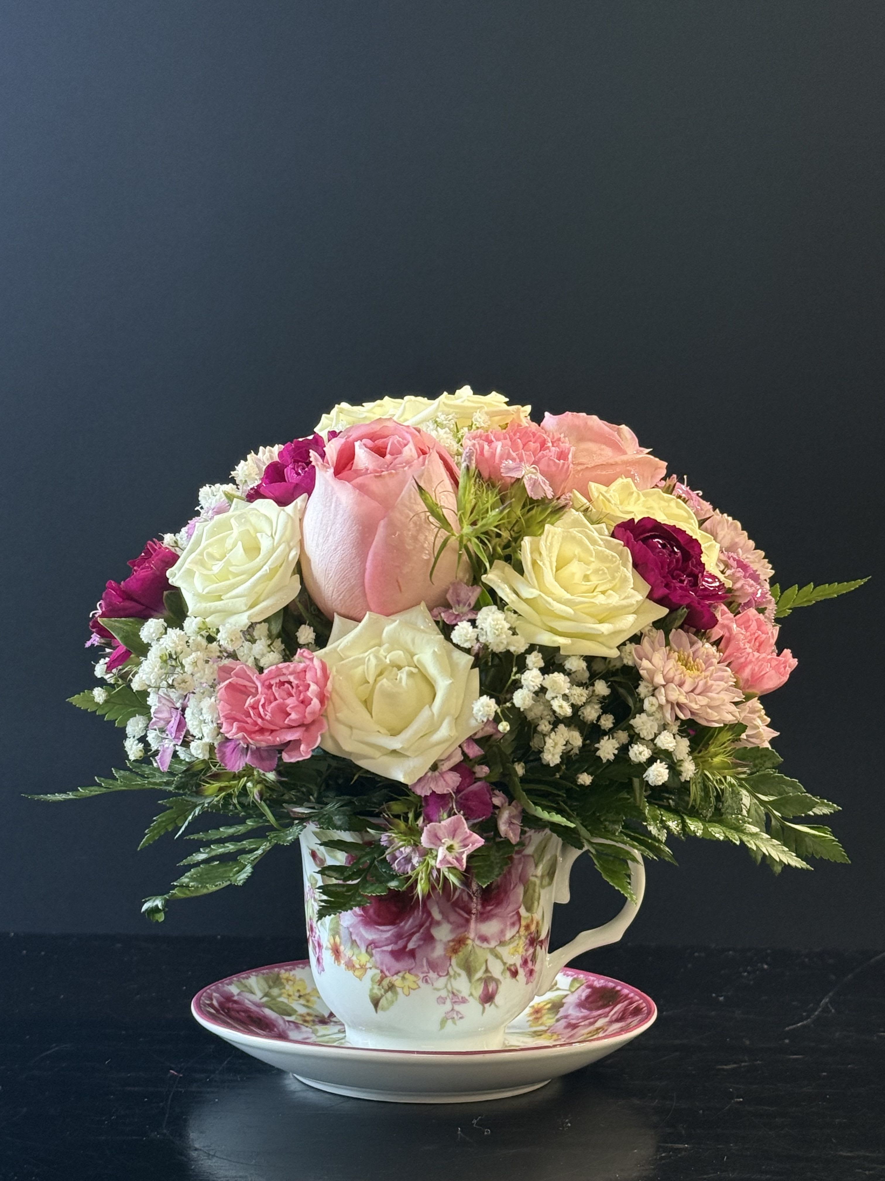 Compact arrangement of pink and white roses and mixed blooms in a floral teacup with matching saucer.
