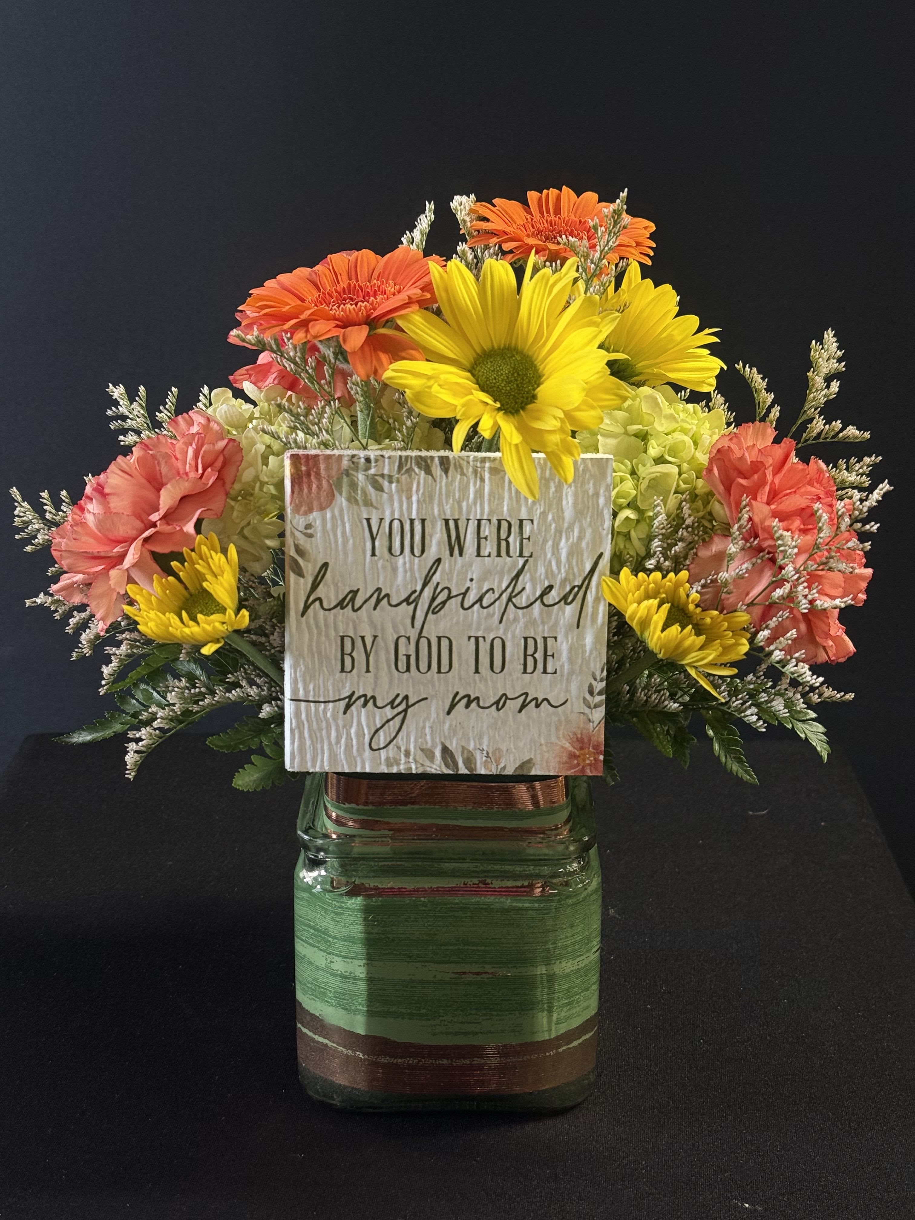 Handpicked By God - Bright orange gerbera, yellow daisies, with orange carnations and pale green hydrangea in a square glass vase, topped with a heartfelt printed sign - warm, cheerful, and perfect for Mother's Day remembrance.