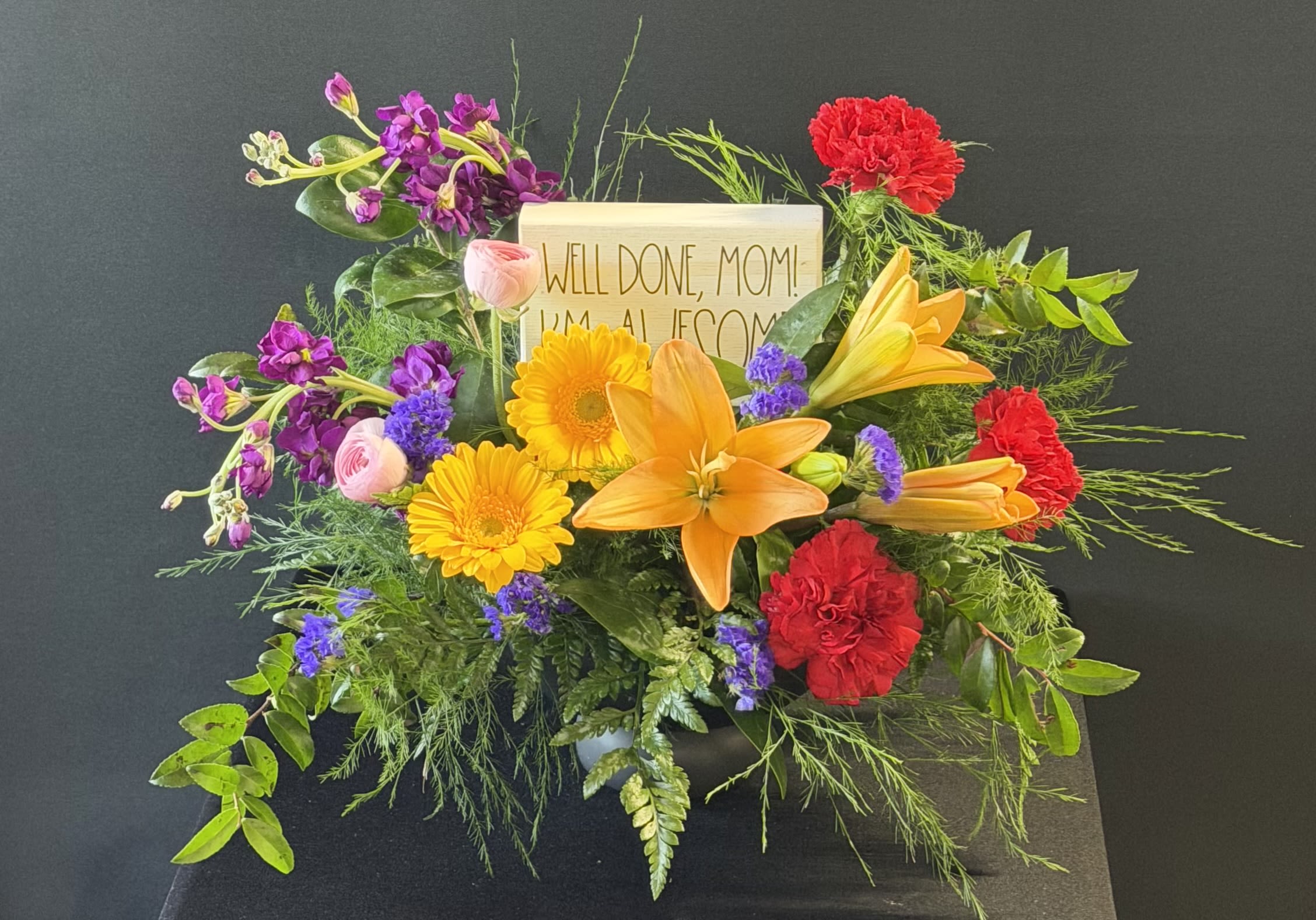 Mother's Day Block with Flowers "Well Done Mom, I'm Awesome" - A burst of celebratory color leads the way: large orange lilies crown a cheerful mixed bouquet made for Mother's Day or any occasion to say "well done." This Mother's Day block with flowers pairs bold yellow gerbera daisies, red carnations, pink blooms and purple accents around airy greenery to create a lively, garden-like silhouette that feels both playful and heartfelt. The included decorative sign - "Well done, Mom! I'm awesome" - gives the arrangement a humorous, personal touch that will make the recipient smile.  Florals are densely arranged for a full, generous look with the orange lily as the focal point and contrasting pops of yellow, purple, pink and red filling the composition. Textures range from the smooth, trumpet shape of lilies to the round faces of gerbera and the ruffled carnations, all anchored by soft filler blooms and sprigs of green for movement. A small card sign is tucked into the back for a supplemental message.  Perfect for a proud mom celebrating a milestone, graduation, or just-for-fun Mother's Day gifting, this bouquet reads as both festive and affectionate - the kind of arrangement that makes her feel seen and celebrated. Order early for best availability; keep blooms in fresh water, trim stems and remove foliage below the waterline to extend vase life.