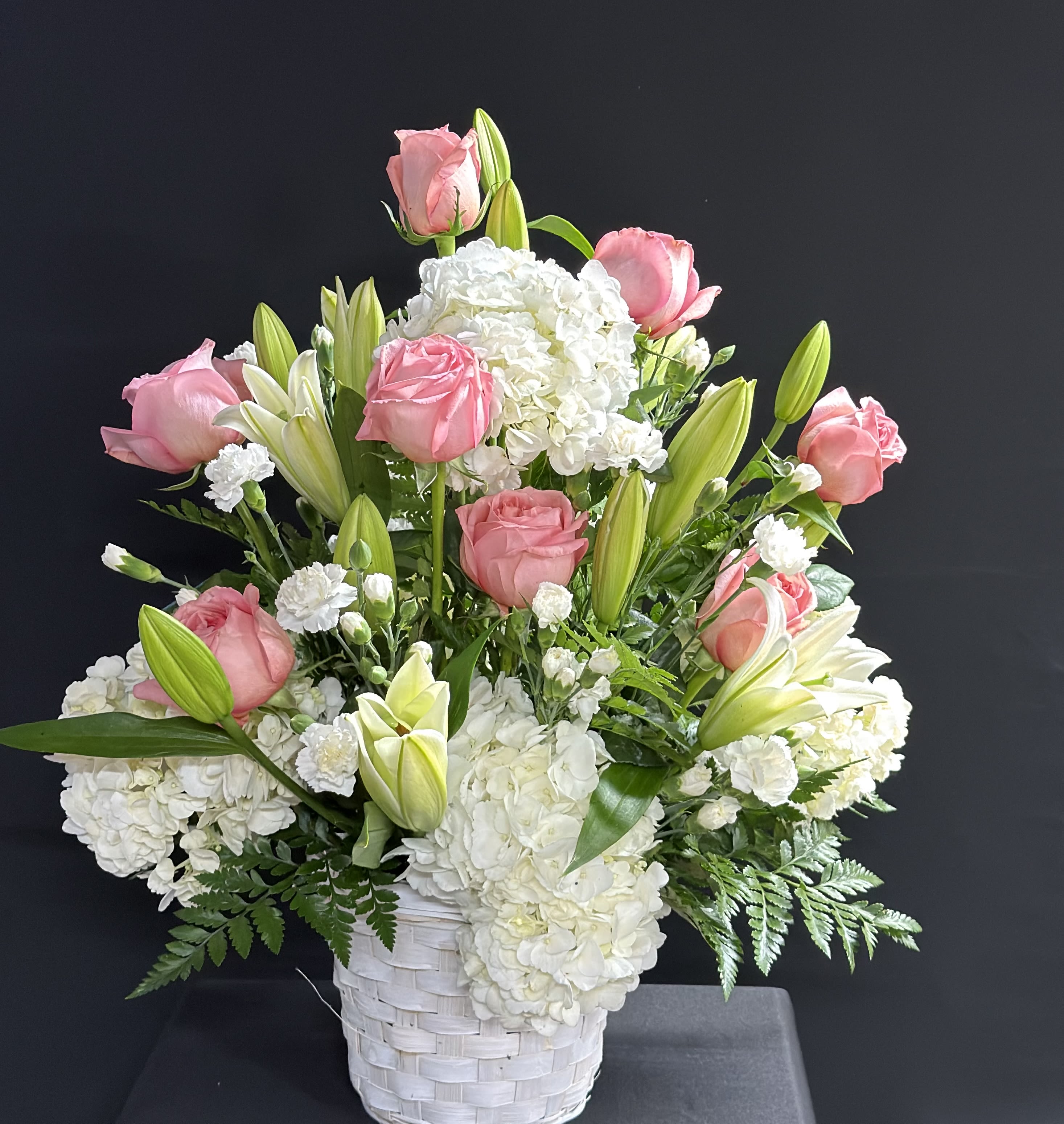 Lily &amp; Rose Basket Floral Arrangement  - Soft pink roses and pale white lilies mingle with lush white hydrangeas and delicate carnations in a white woven basket, creating a soft, elegant floral gift perfect for birthdays, anniversaries, or a thoughtful everyday surprise. This lily &amp; rose basket floral arrangement reads as classic and gently formal-she'll feel genuinely celebrated by the fresh, romantic palette and the graceful height of blooms rising above a full, rounded base.  The design is full and layered: large hydrangea clusters form a dense foundation while roses, lilies, and carnations provide texture and vertical interest for a balanced silhouette. Presented in a tasteful white woven basket, the arrangement brings a fresh, classic look to mantels, entry tables, or bedside displays; colors are white, pink, and green for a timeless appeal. Ideal for sending comfort, congratulations, or thanks, this piece arrives ready to display-no extra vase needed-and keeps its airy, garden-fresh presence wherever it's placed.