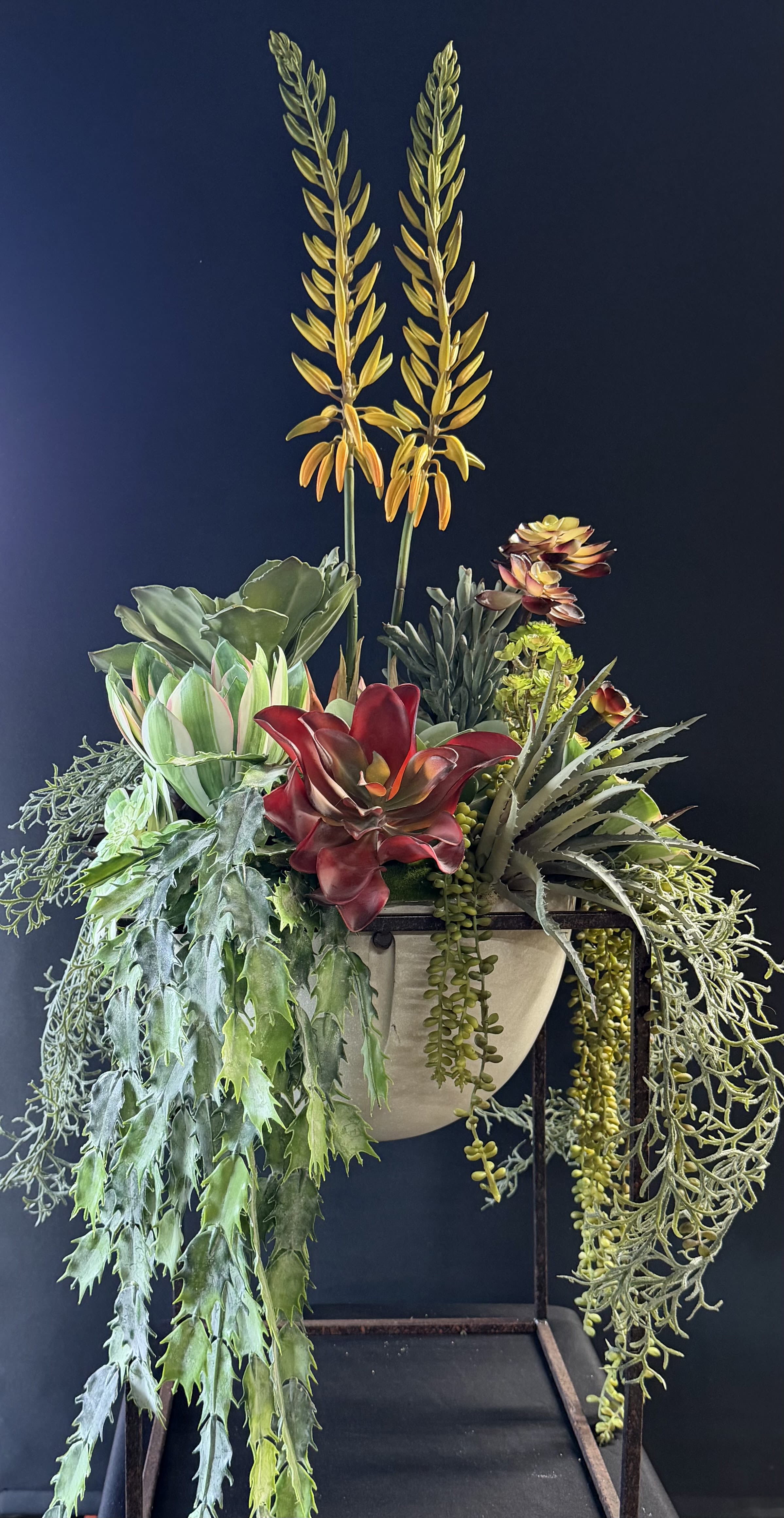 Faux Succulent Planter - A sculptural standing succulent planter that brings a modern desert-garden vibe to everyday decor or a housewarming gift. This planter arrangement-perfect for gifting a friend who loves low-maintenance style or for adding texture to a living room or office-features a bold centerpiece of deep red and pale green rosettes with spiky gray-green forms and two tall yellow-orange vertical spikes that catch the eye.  The dense mix of succulents and trailing foliage is arranged in a compact container set on a clean metal stand for display; cascading plants spill well below the planter for dramatic movement and a layered, textural look. Greens, reds, yellows, and oranges play together for warmth and contrast, creating a piece that reads like living art rather than a simple pot of plants.  Ideal for the person who wants the look of a lush indoor garden without the upkeep, this faux succulent planter stays vibrant without watering or special care-an easy way to make someone feel seen and celebrated. Its wall-mounted design fits small spaces, entryways, and galleries; choose a spot with bright, indirect light to keep the display looking its best.