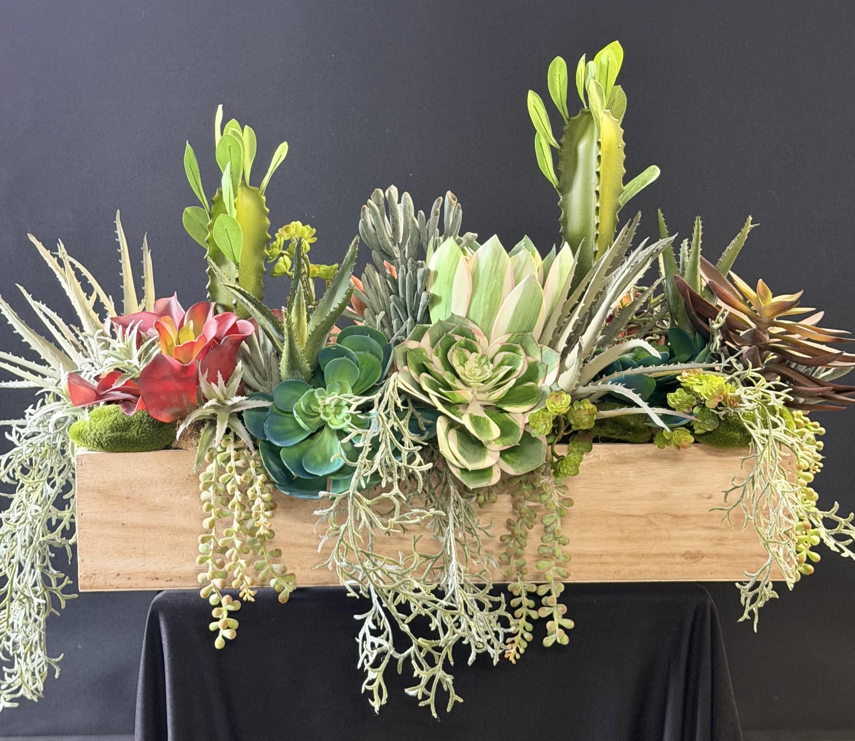 Faux Succulent Box - A sculptural mix of faux succulents and cactus-like plants arranged in a long rectangular wooden planter-perfect as a modern housewarming gift, anniversary accent, or low-maintenance birthday surprise. The piece reads like a carefully curated mini desert garden, combining upright spiky forms, rounded rosettes, and trailing strands so the person you're thinking of will feel seen by a design that's both lively and grounded.  Greens dominate the palette-soft chartreuse, silvery sage, and deeper forest tones-punctuated by pops of pink, red, yellow, blue and pale cream for contrast and texture. The dense composition creates layered visual interest from every angle, with several trailing plants that spill naturally over the planter's edge. Housed in a rectangular wooden planter, this arrangement brings enduring, natural style to desks, entry tables, mantels, or windowsills without watering or upkeep.  Ideal for Father's Day, Spring or Summer décor, anniversaries, sympathy and funeral settings where a lasting, tasteful presence is preferred, or as a thoughtful gift for Mother's Day, Valentine's Day, Boss's Day, or a new home. Choose from size options at checkout; care is simple-dust occasionally and keep out of prolonged direct moisture to preserve the lifelike finish.