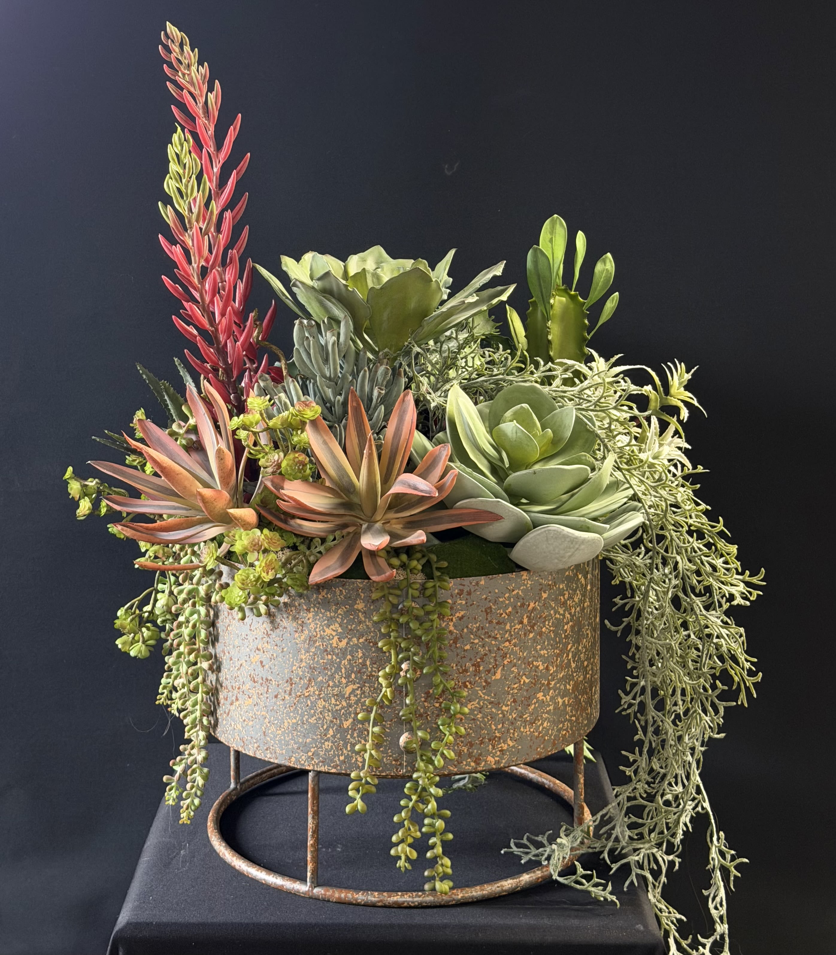 Faux Succulent Metal Plant Stand - A sculptural faux succulent arrangement that reads like modern rustic home decor-perfect as a housewarming gift, thank-you, or just-because surprise. This flower arrangement features a weathered round metal planter set on a matching stand, so it functions as both a decorative accent and an effortless way to bring sculptural green style into any room. The look is contemporary with desert-inspired charm, ideal for someone who loves low-maintenance, long-lasting greenery.  The mix includes dense faux succulents and textured trailing plants in a soft green palette, punctuated by coral-pink and orange rosettes and a vivid pink-red vertical accent that adds height and drama. Cascading strands spill over the sides for a lush, layered silhouette, while the rust-toned, weathered metal planter and round cylindrical form give the piece an authentic, aged finish. No watering or sunlight requirements-this arrangement keeps its shape and color year-round.  Give a recipient a display that feels both intentional and effortless: it makes a thoughtful housewarming centerpiece, a modern thank-you, or a welcoming accent for a new office. Sized to sit on the floor or a low table thanks to its sturdy metal stand, it arrives ready to style-no care needed beyond occasional dusting.