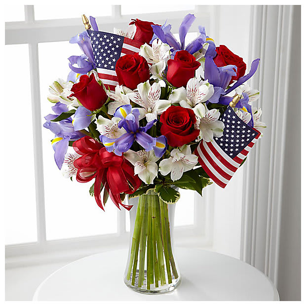 Unity Bouquet - The Unity Bouquet sparks the hearts of all Americans with its patriotic beauty and dazzling color. Bright red roses mingle with blue iris arranged amongst white Peruvian lilies and assorted greens. Accented with two American Flags and a red, white and blue ribbon, this stunning bouquet arrives arranged in a clear glass vase to creae a gorgeous way to celebrate our Independence Day.