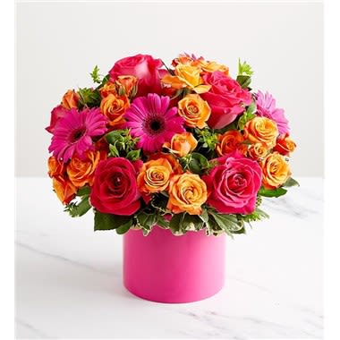 Sunset Radiance Bouquet - This bouquet captures the vivid blend of hot pink, bright orange, and golden yellow flowers, evoking the warmth and energy of a vibrant sunset. All-around arrangement of hot-pink roses, orange spray roses, and hot-pink mini gerbera daisies, designed in a modern 6 inch ceramic hot pink cylinder, complemented with assorted greenery.  Large arrangement measures approximately 17”H x 13”W Medium arrangement measures approximately 16”H x 12”W Small arrangement measures approximately 15”H x 11”W Our Designers hand-design each arrangement, so flowers varieties, colors, and container may vary due to local availability.