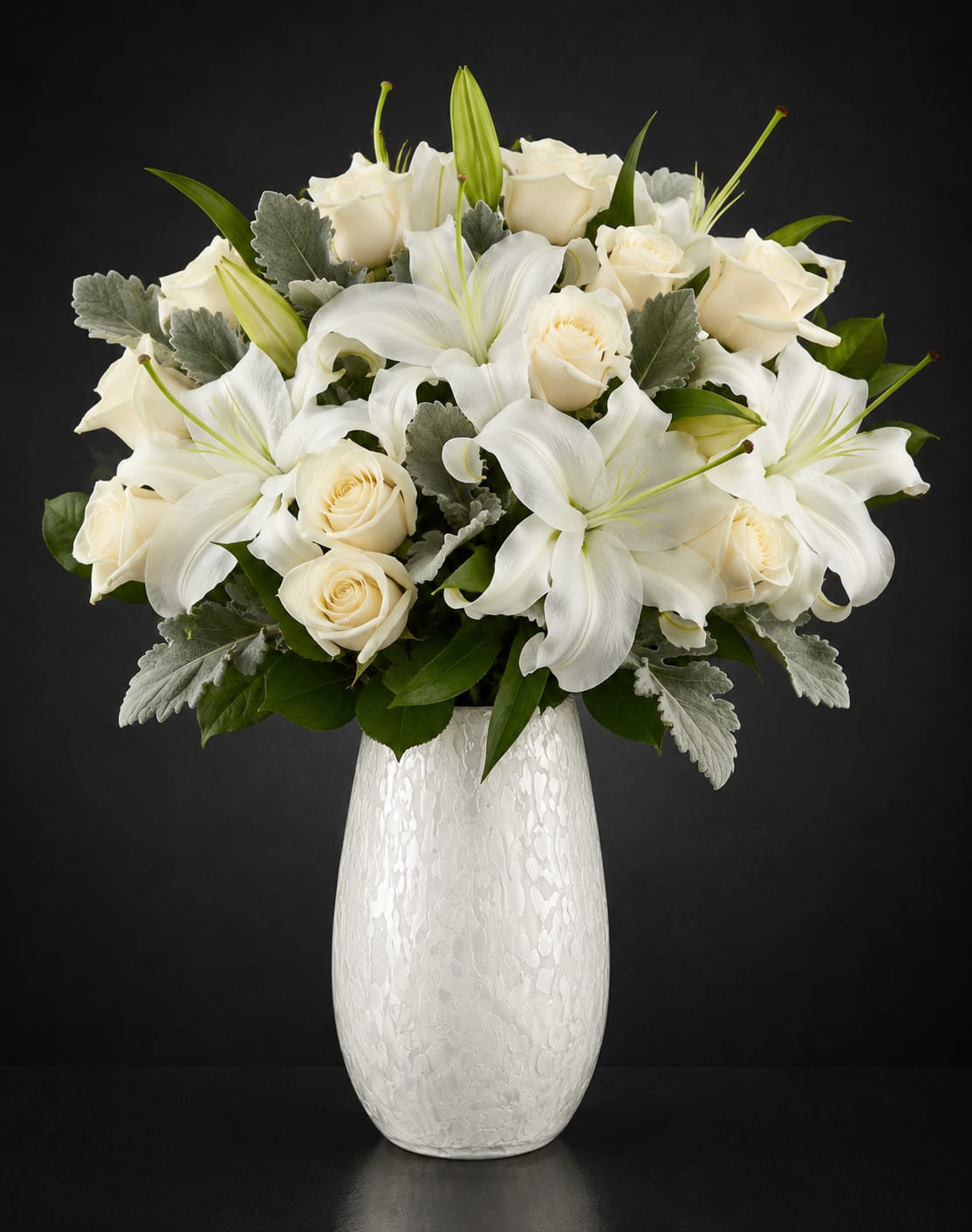 Pure Opulence Luxury Bouquet - A refined mix of ivory roses and broad white lilies creates a tall, luxurious bouquet that reads as pure elegance-perfect as an anniversary bouquet or a tasteful sympathy arrangement. The restrained palette of soft cream petals and bright lily faces, lifted by muted silver-green foliage, gives the person you're thinking of a calm, comforting impression that also suits weddings, birthdays, Mother's Day, Valentine's Day, holiday gifting, or a thoughtful "just because" moment.  This arrangement is gathered into a full, rounded design with a taller profile to showcase the lilies' dramatic faces and the roses' creamy texture. The stems sit in a textured white ceramic vase that adds subtle pattern and a modern, high-end feel while keeping the overall look serene and formal. Sized to make a meaningful statement without overwhelming a room, it's an elegant choice for anyone who appreciates classic whites and soft greenery-easy to display at home, at a ceremony, or beside a funeral service program.