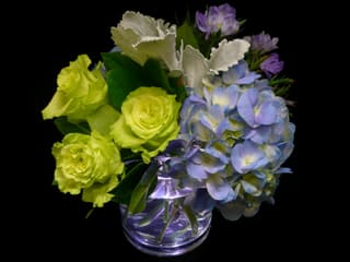 Compact arrangement of green roses and blue hydrangeas in a clear glass vase on a black background.