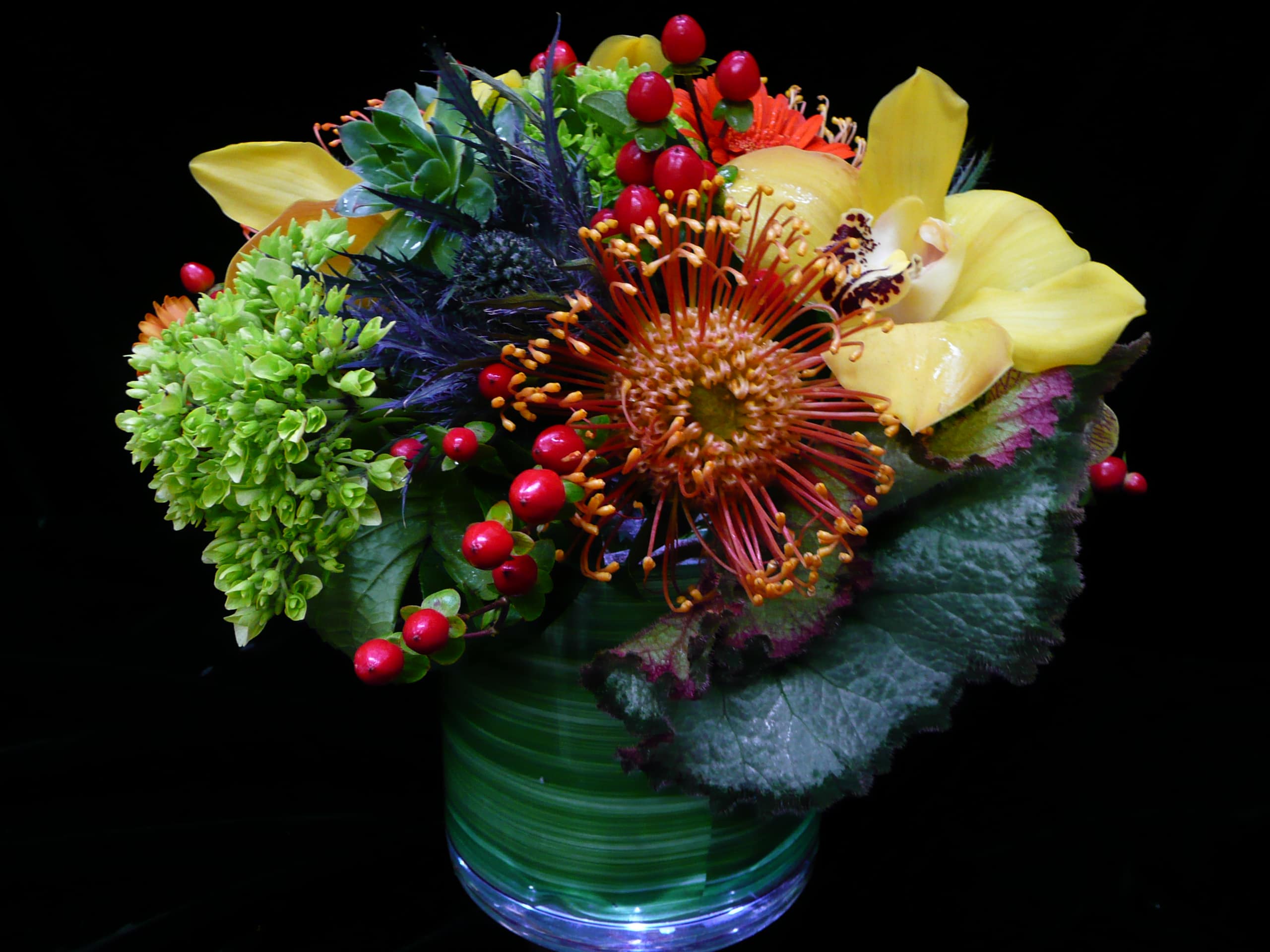 Low arrangement of green hydrangea, orange protea, yellow orchid, succulents, and red berries in a glass vase