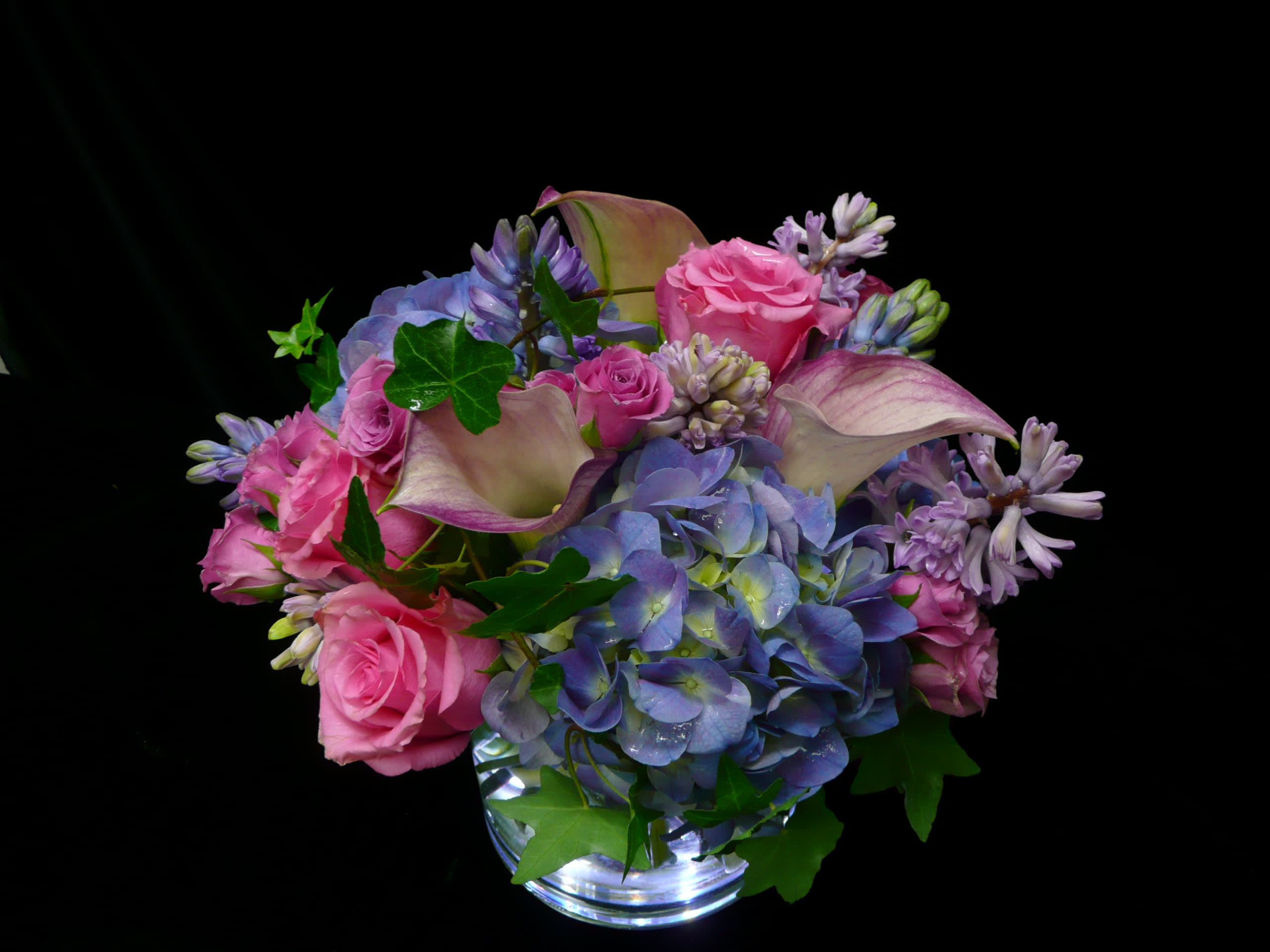 Low arrangement of pink roses, blue hydrangeas, and pale calla lilies in a clear glass vase.