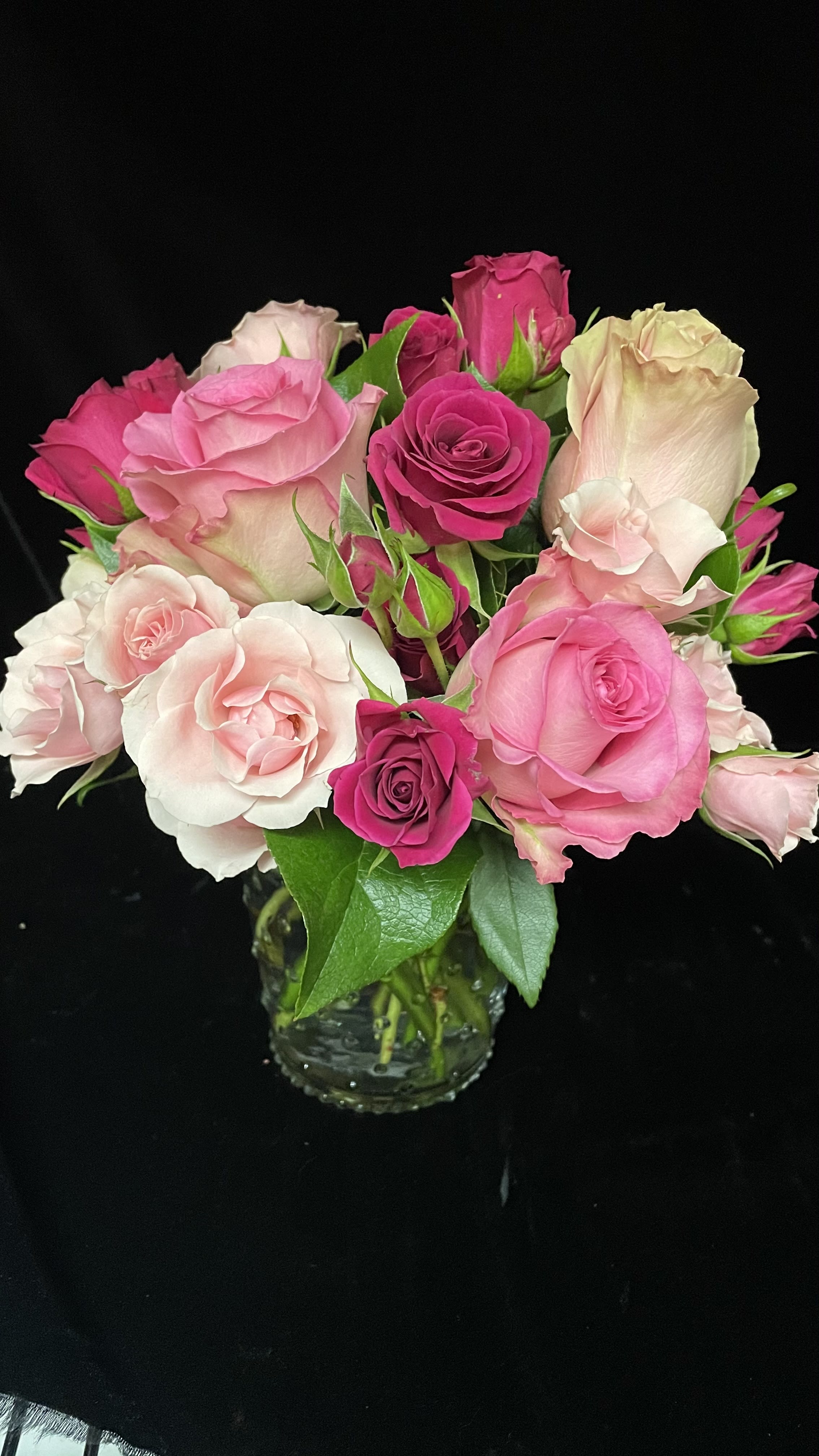 Pink and blush roses arranged tightly in a small clear glass vase against a black background