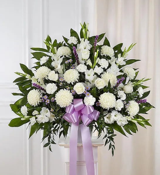 Large fan-shaped arrangement of white flowers with purple accents and a lavender bow on a white pedestal
