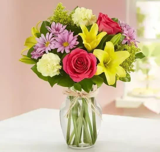 Mixed bouquet of pink roses, yellow lilies, purple daisies, and cream carnations in a clear glass vase