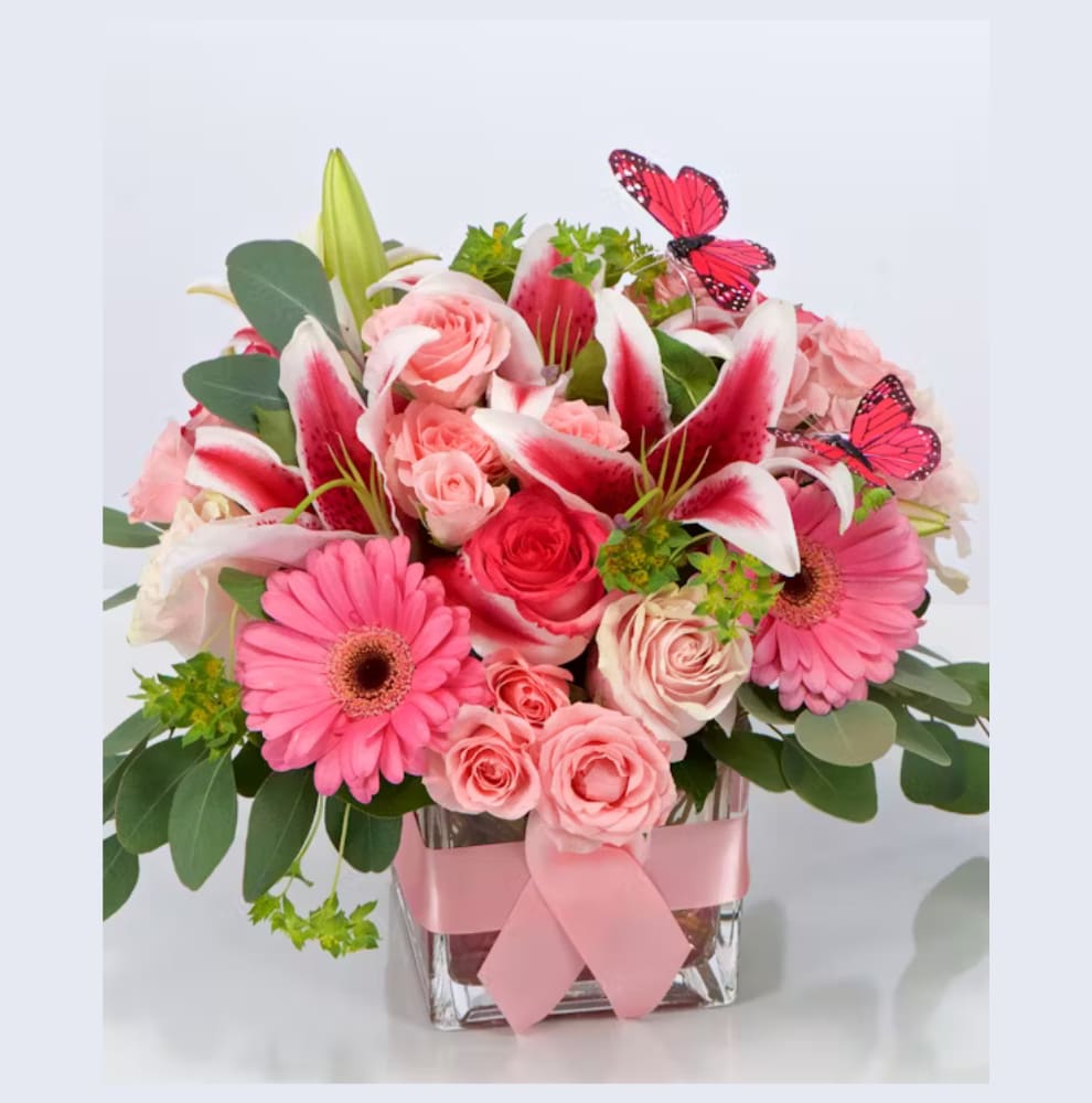 Pink lilies, roses, and gerbera daisies in a glass cube vase with pink ribbon and butterfly accents