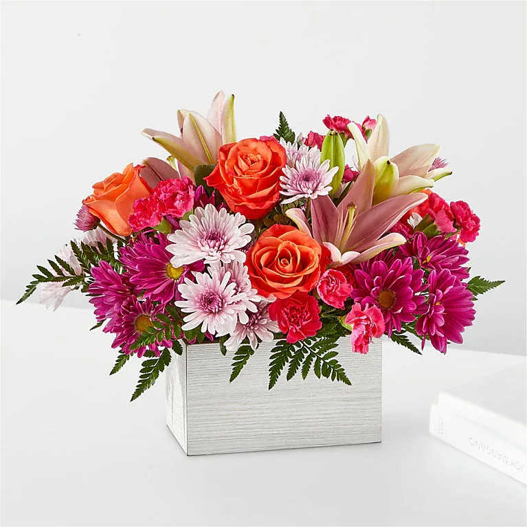 Low rectangular arrangement of orange roses, pink lilies, and mixed pink and white mums in a white box vase