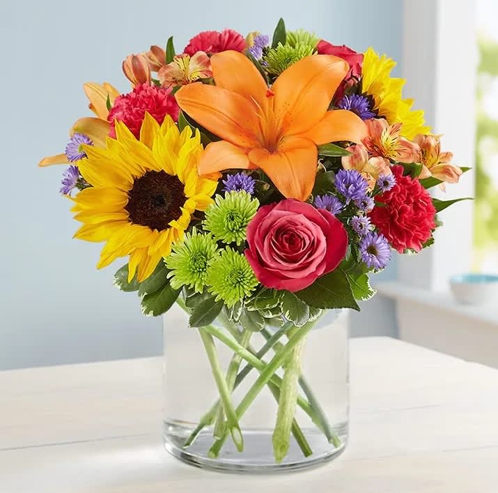 Floral Embrace - Like a warm embrace, our vibrant bouquet delivers your sentiments to someone special. A rich gathering of yellow and orange blooms, with pops of bright pink and purple, it's more than a gift - it's a way to express how you feel inside. All-around arrangement of hot pink roses and carnations; orange Asiatic lilies and Peruvian lilies (alstroemeria); yellow sunflowers or similar blooms; lavender stock; purple monte casino; green Athos poms; accented with assorted greenery Artistically designed in a glass vase Large arrangement does not include carnations